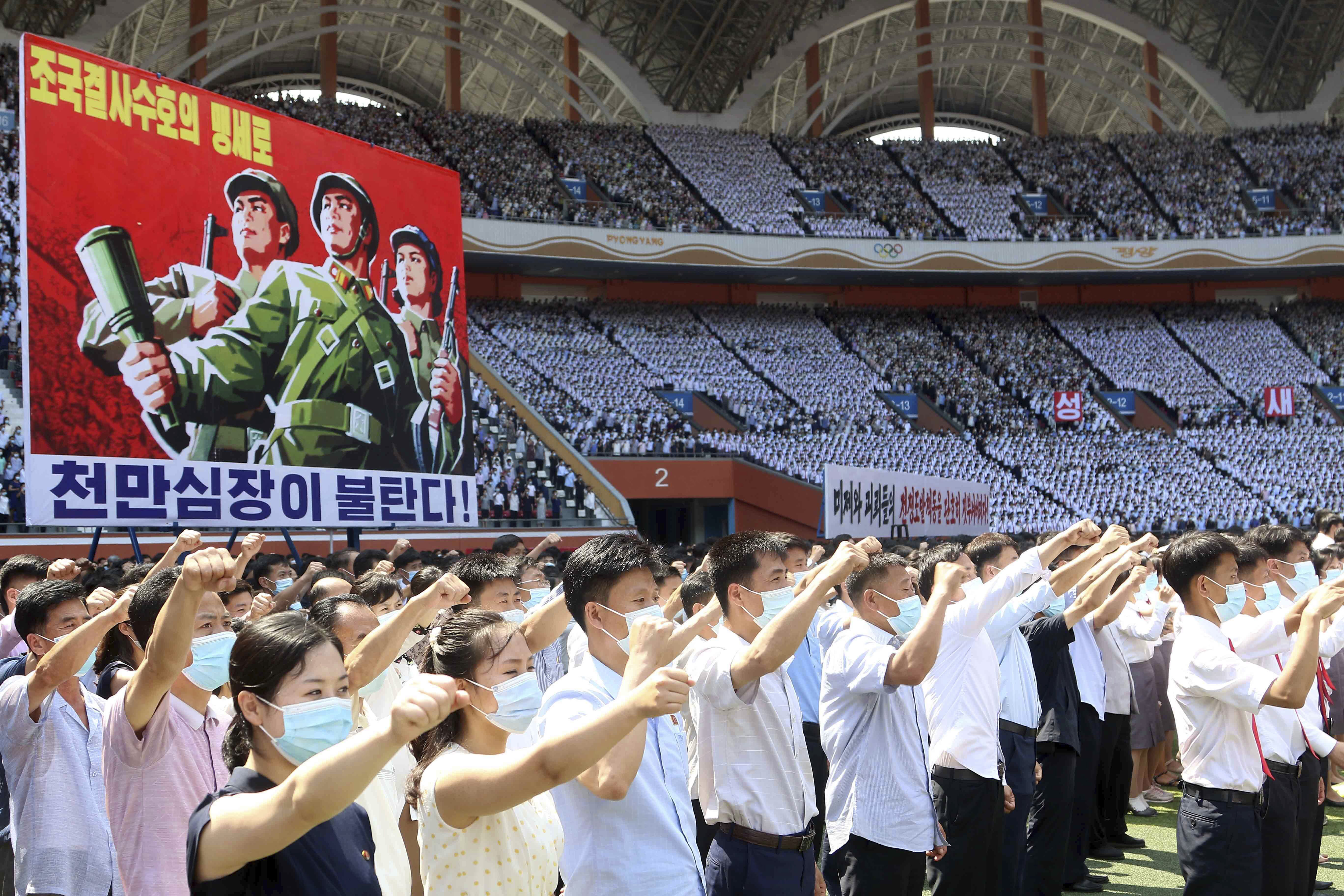 A crowd of North Koreans punch the air at a rally to denounce the US. A poster behind them shows an illustration of three armed North Korean soldiers against a red background. The slogan reads: Tens of millions of people pledge to defy death for defending the country!