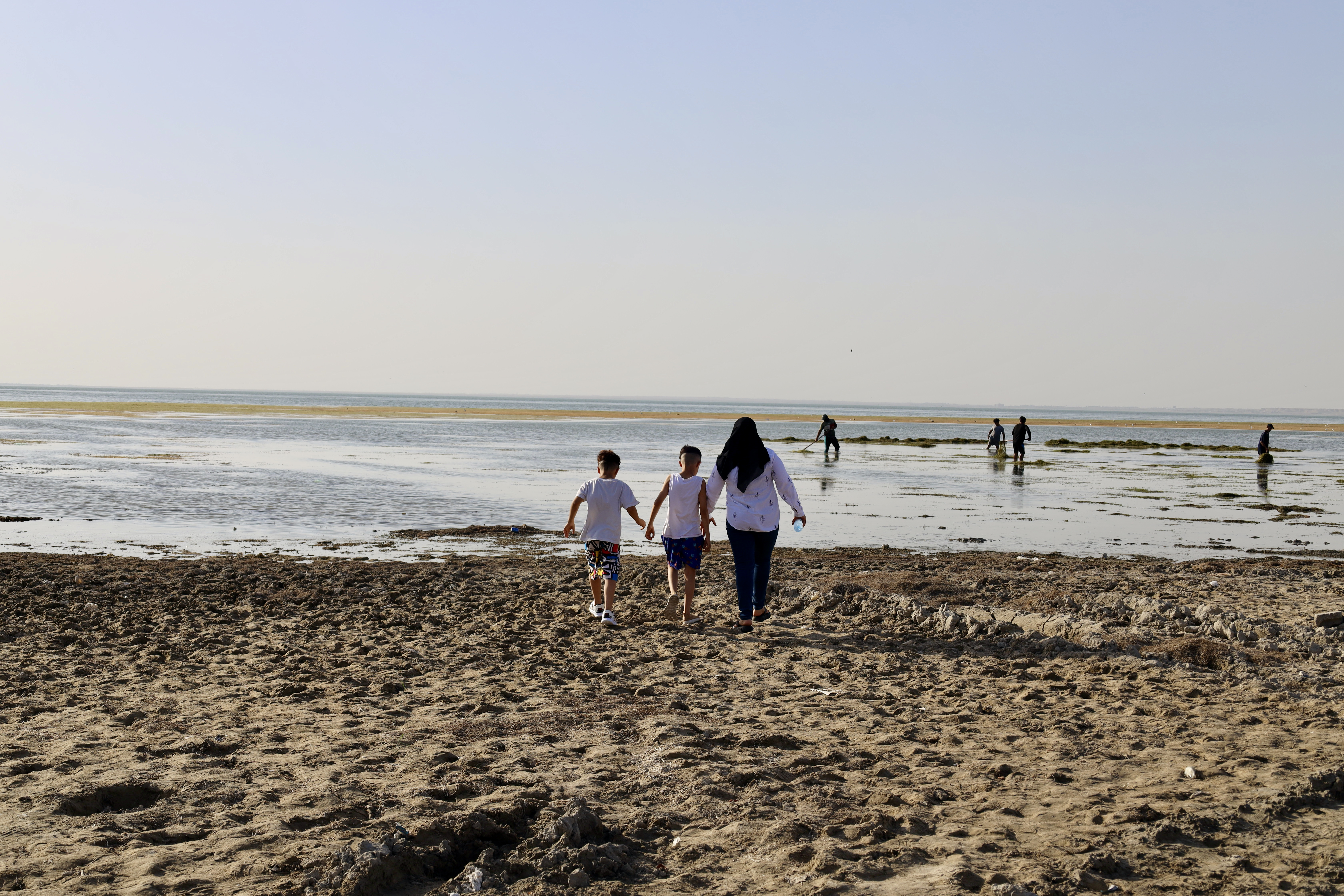 Noor Haddi, 35, and her two young sons venturing towards the distant water of the Habbaniyah Lake resort. In front of them, day labourers earn a  daily wage by cleaning natural debris and trash from the water.