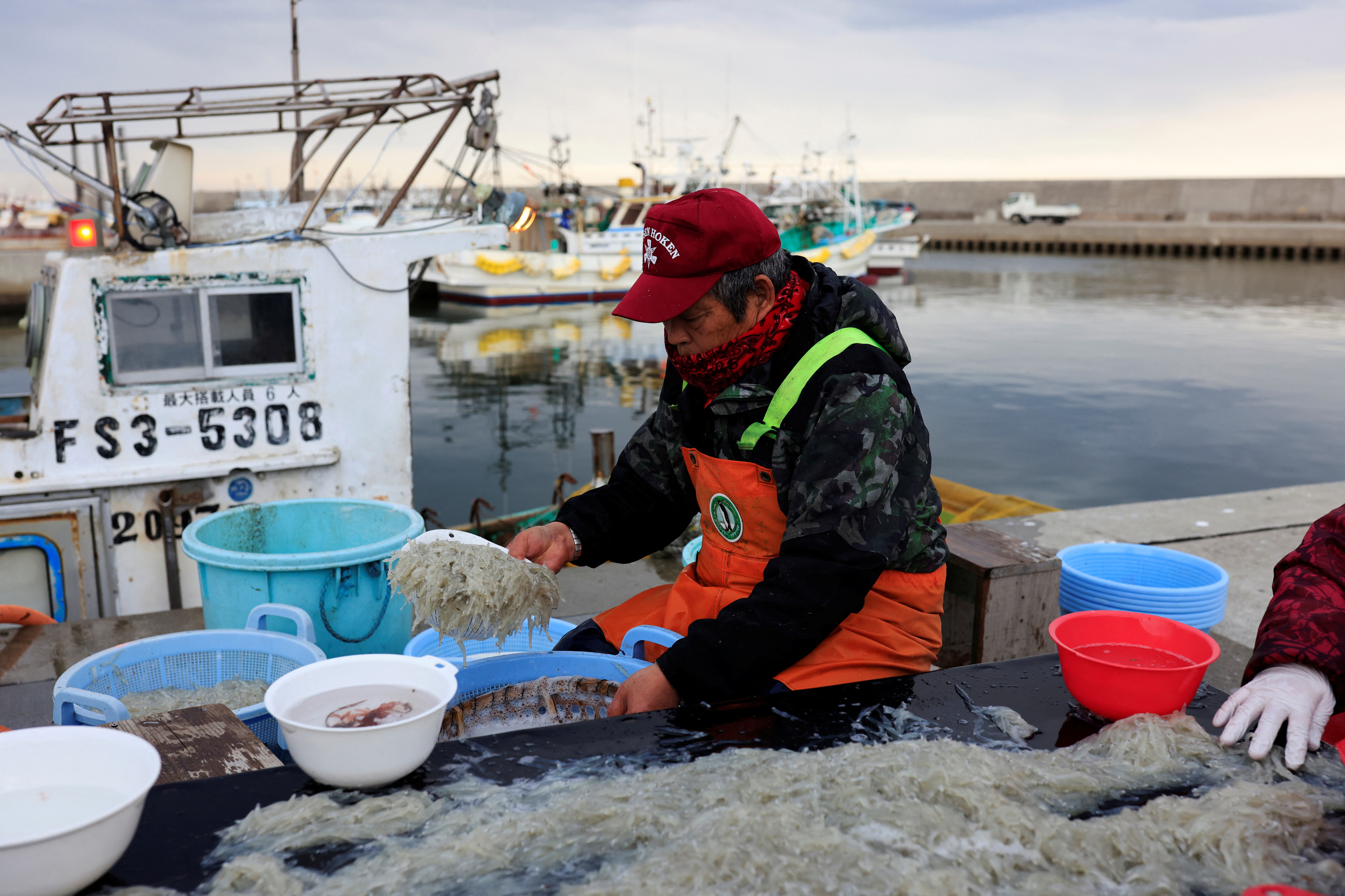 A fisherman sitting on the quayside washing fish. He is wearing wet weather gear and a red cap. Fishing boats are behind him. The tiny fish are piled up in front of him.