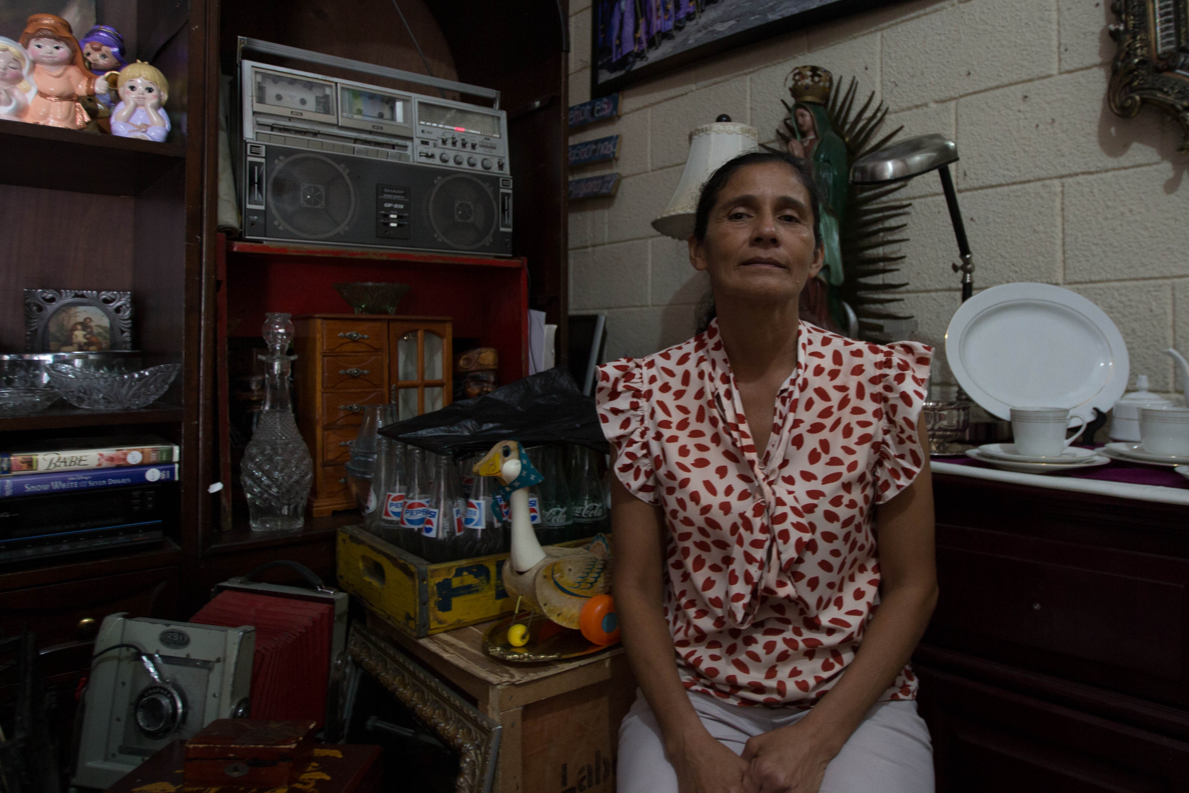 A woman in a dotted patterned shirt sits in a shop with items for sale: glass bottles, plates, teacups, glass drink bottles and porcelain figurines.