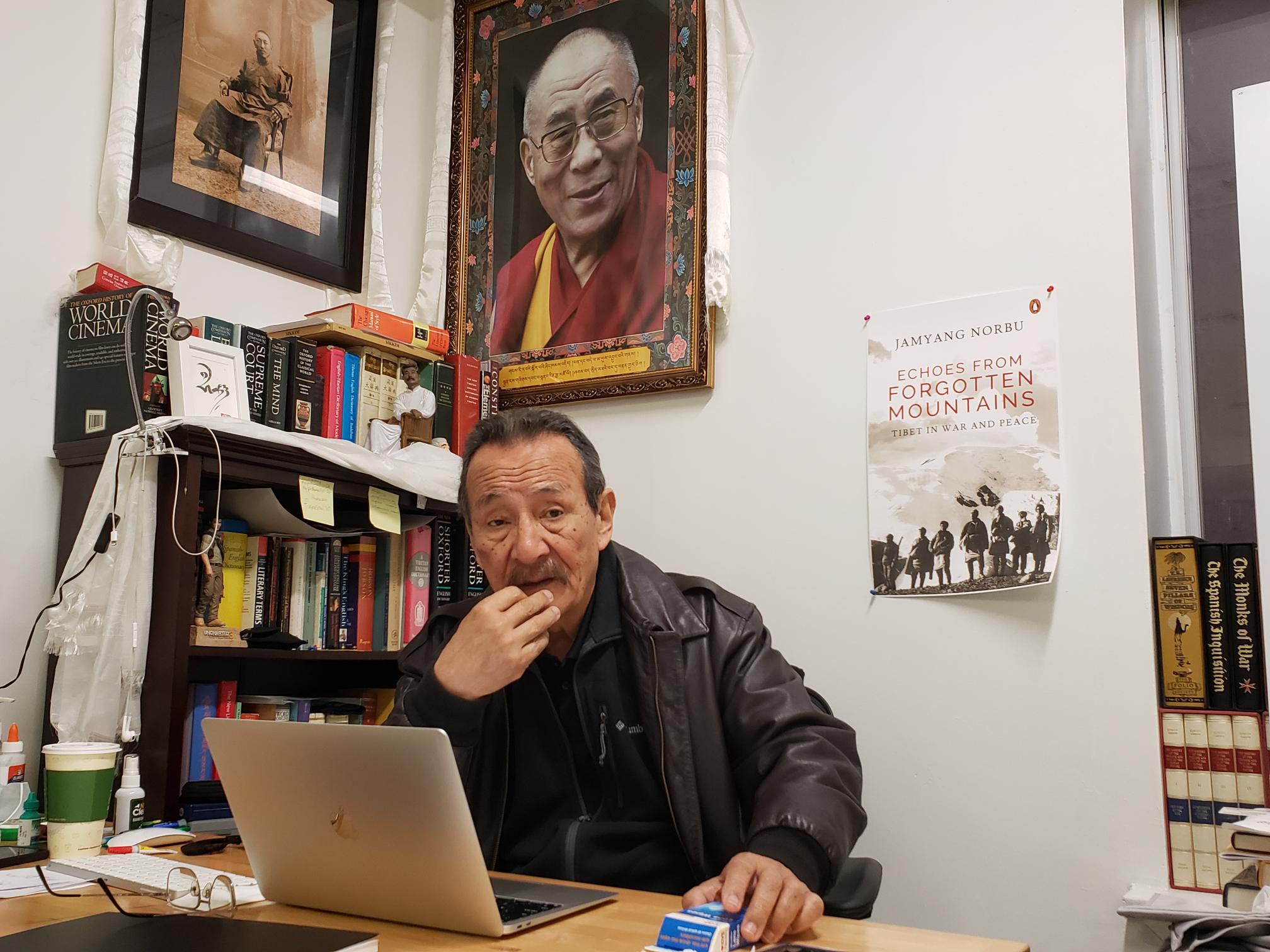 Jamyang Norbu at his desk, He has a computer open in front of him. There is a book shelf to his right and a large portrait of the Dalai Lama on the wall behind him. A mock-up of the front cover of his new book is also on the wall