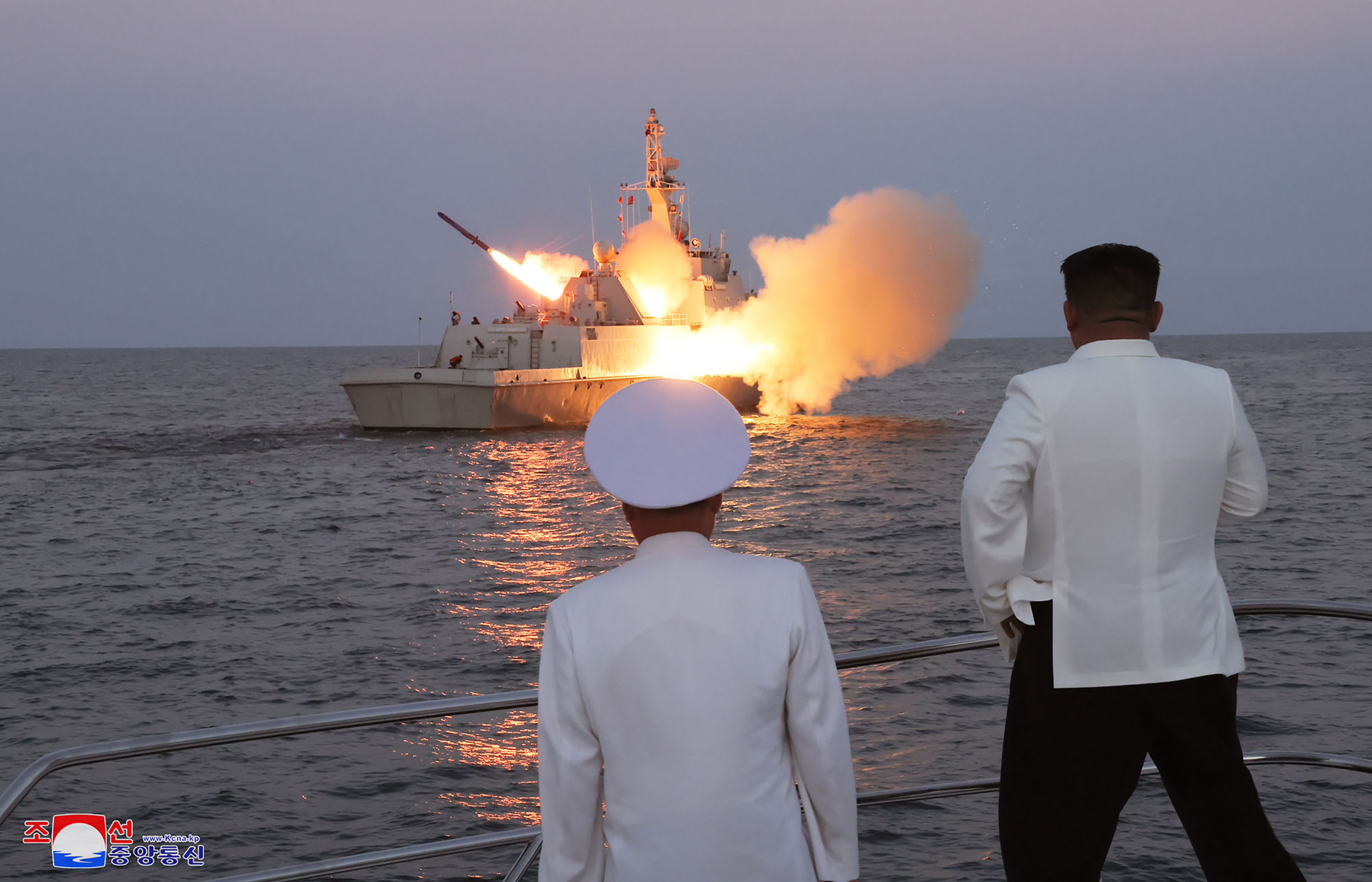 A missile is launched from a boat in a blaze of flames. Kim Jong Un and a navy officer are watching from the quay. They have their backs to the camera.