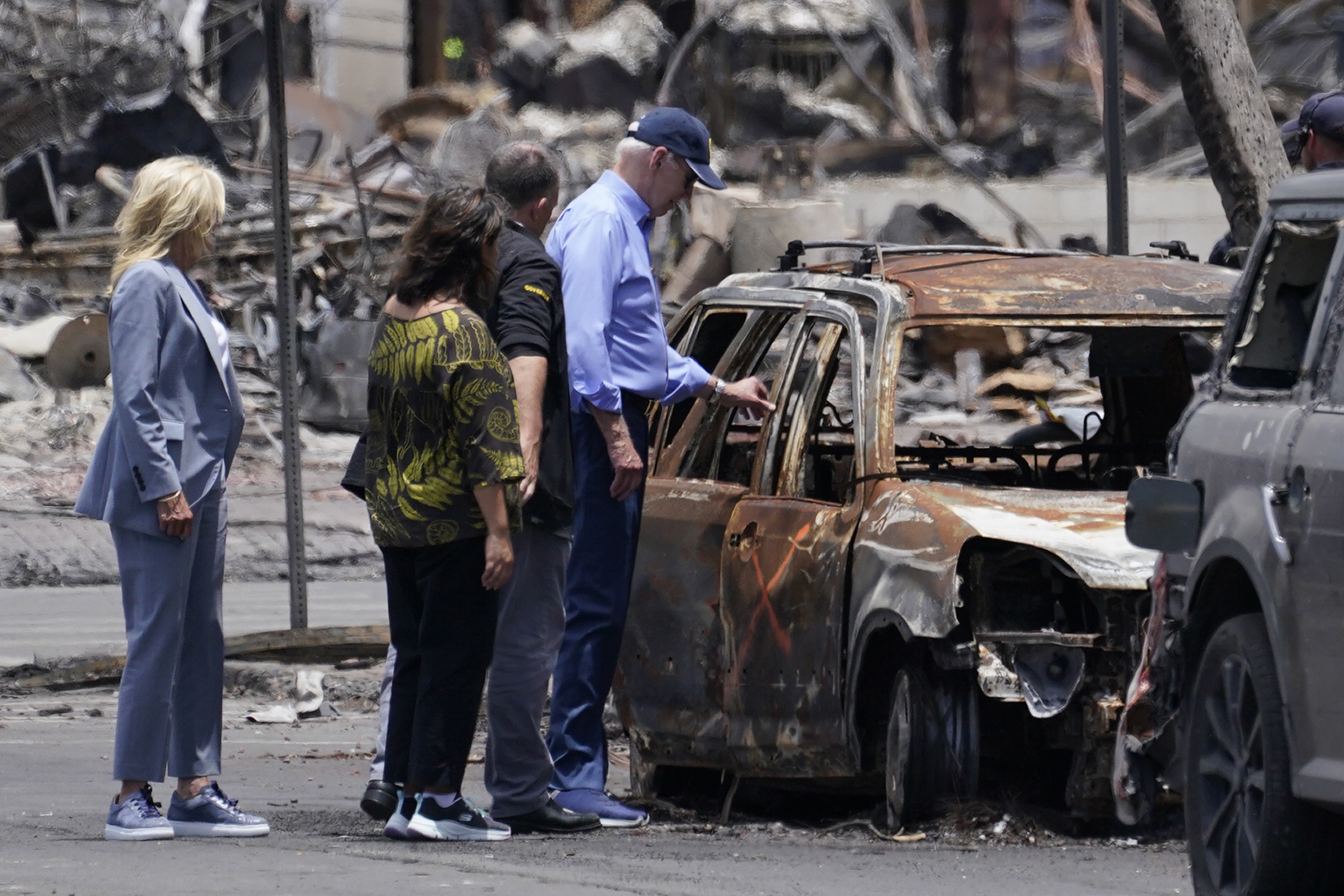 Joe Biden reaches out to touch a burned car, its windows gone and its exterior a mottle of grey and rust colors. Behind him are members of the community and his wife, Jill Biden.