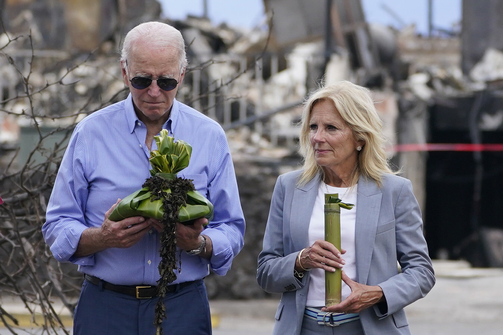 Joe and Jill Biden hold ceremonial items — intricately folded palm fronds and bamboo stalk — as part of a blessing ceremony in Lahaina. The destruction wrought from the fire is visible behind them.