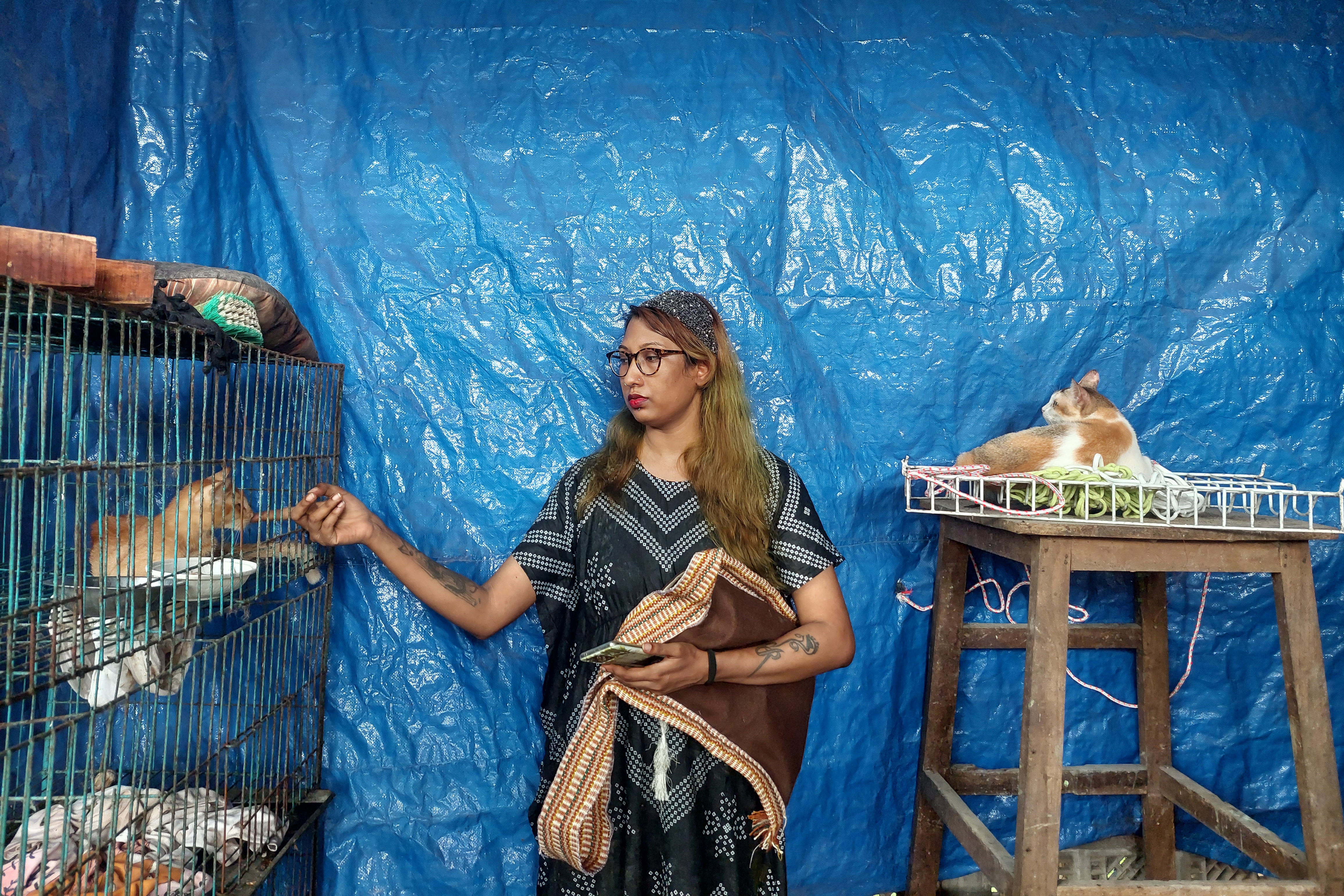 A photo of a woman standing in between a cage with cats on the left and a tall table with a cat sitting on top on the right, while she plays with the cat in the cage.