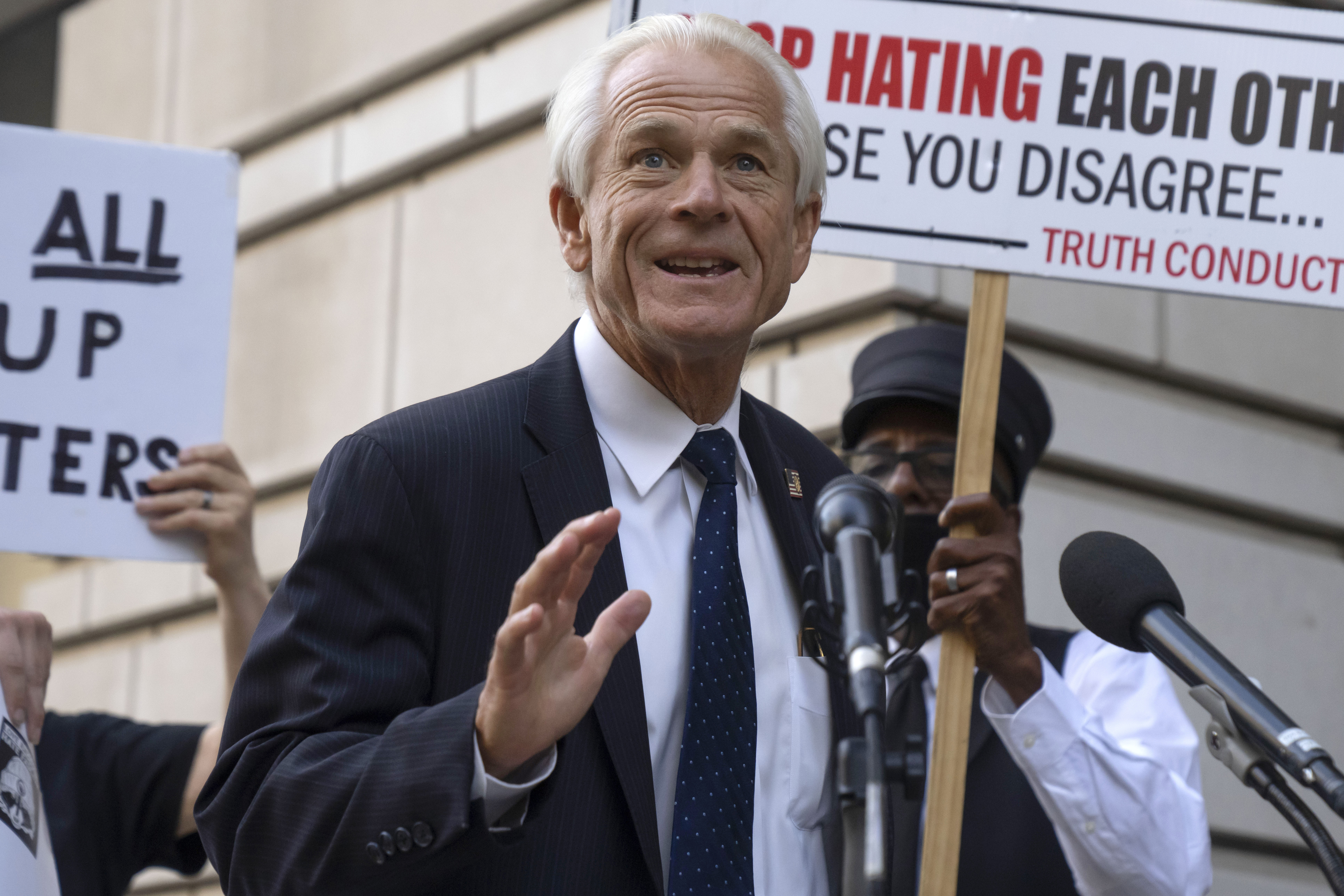 Former White House trade adviser Peter Navarro speaks to the media as he departs federal court, Tuesday, Sept. 5, 2023, in Washington. Navarro was convicted Thursday, Sept. 7, of contempt of Congress charges filed after he was accused of refusing to cooperate with a congressional investigation into the Jan. 6, 2021, attack on the U.S. Capitol. Dressed in a suit and tie, Navarro waves — but behind him are protesters carrying signs.
