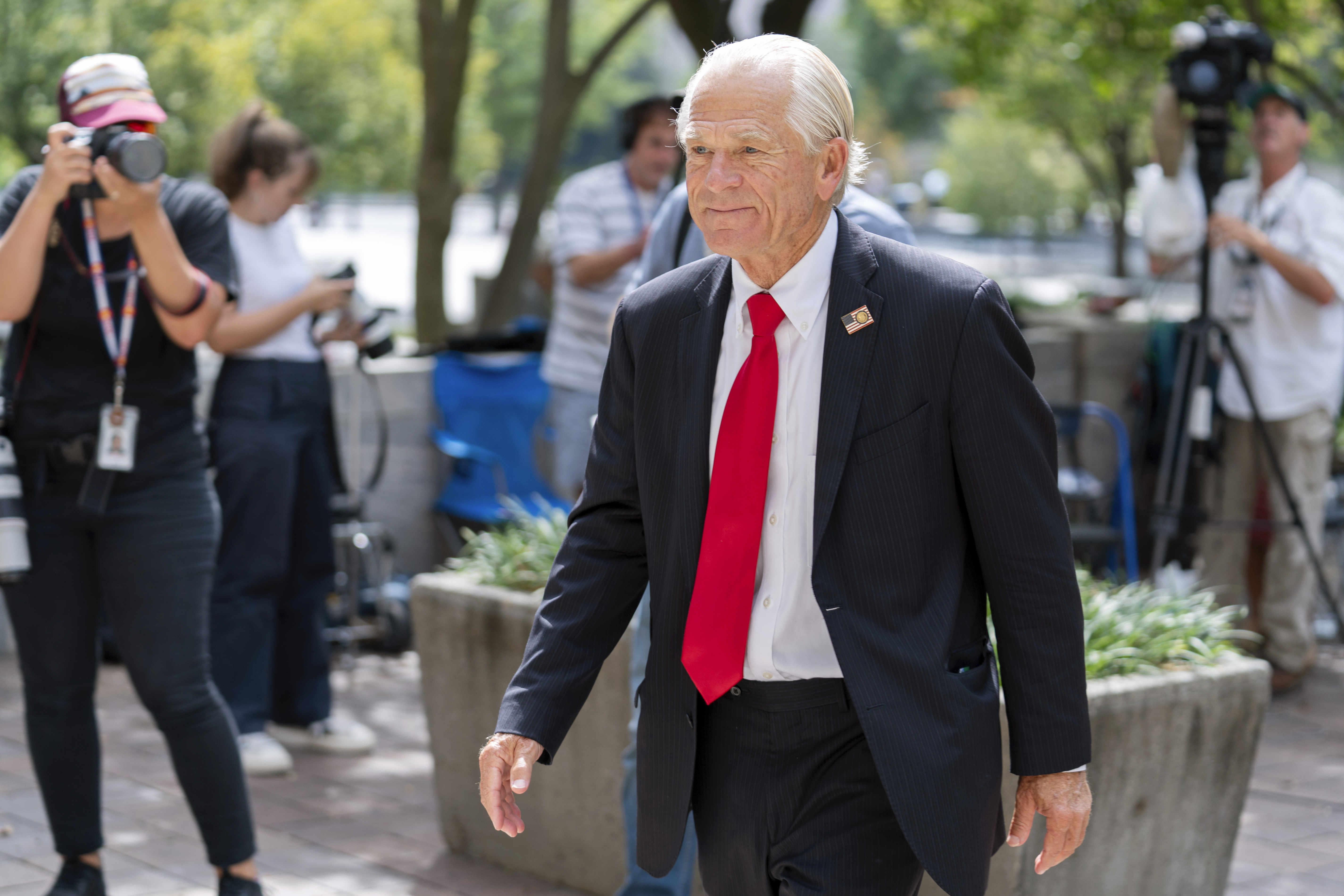 A white-haired man in a dark suit and red tie walks through an outdoor area with leafy potted trees and reporters standing nearby.