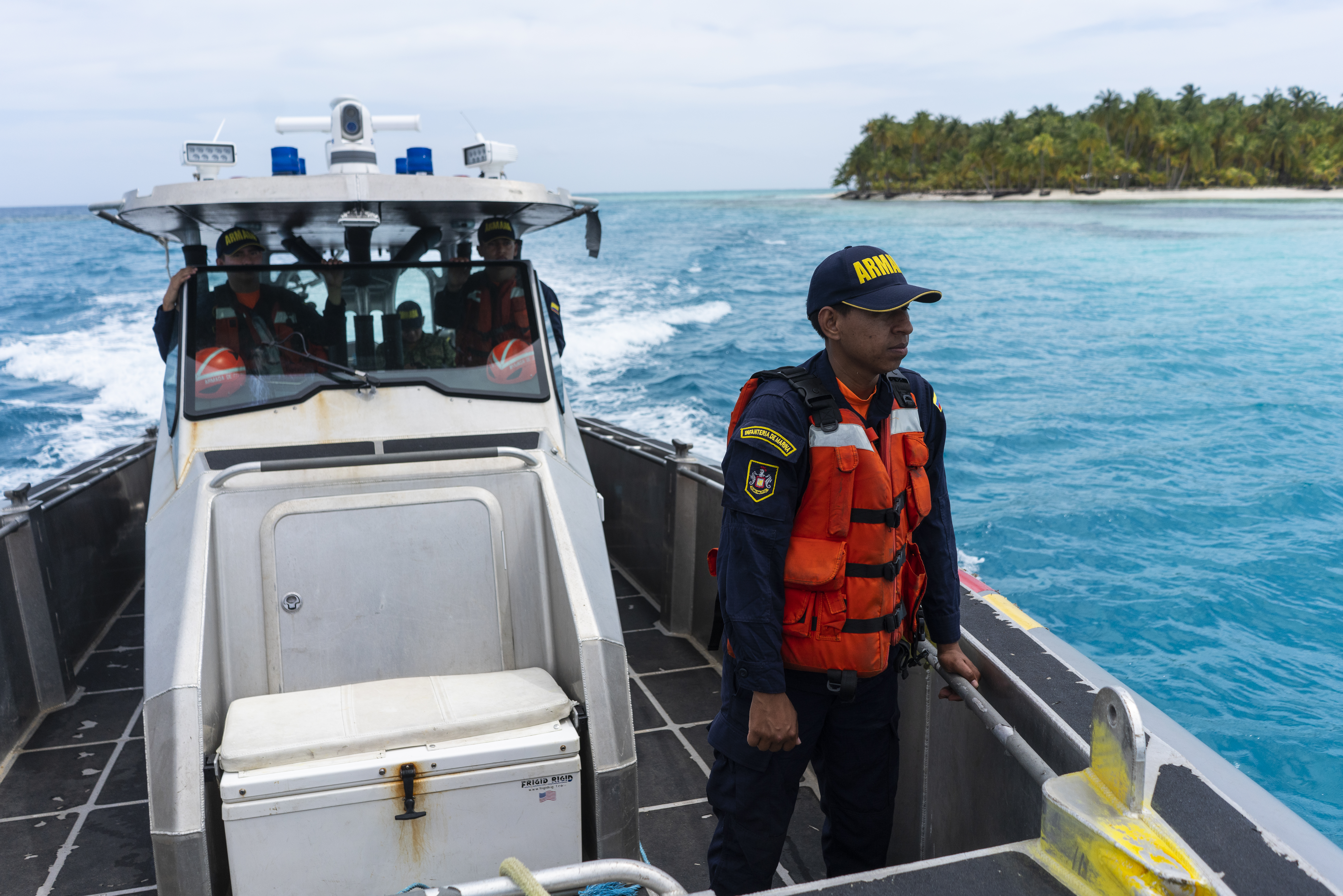A Navy official, dressed in a ballcap, a long-sleeved uniform and an orange life vest stands aboard a boat that plies its way through the blue Caribbean waters.
