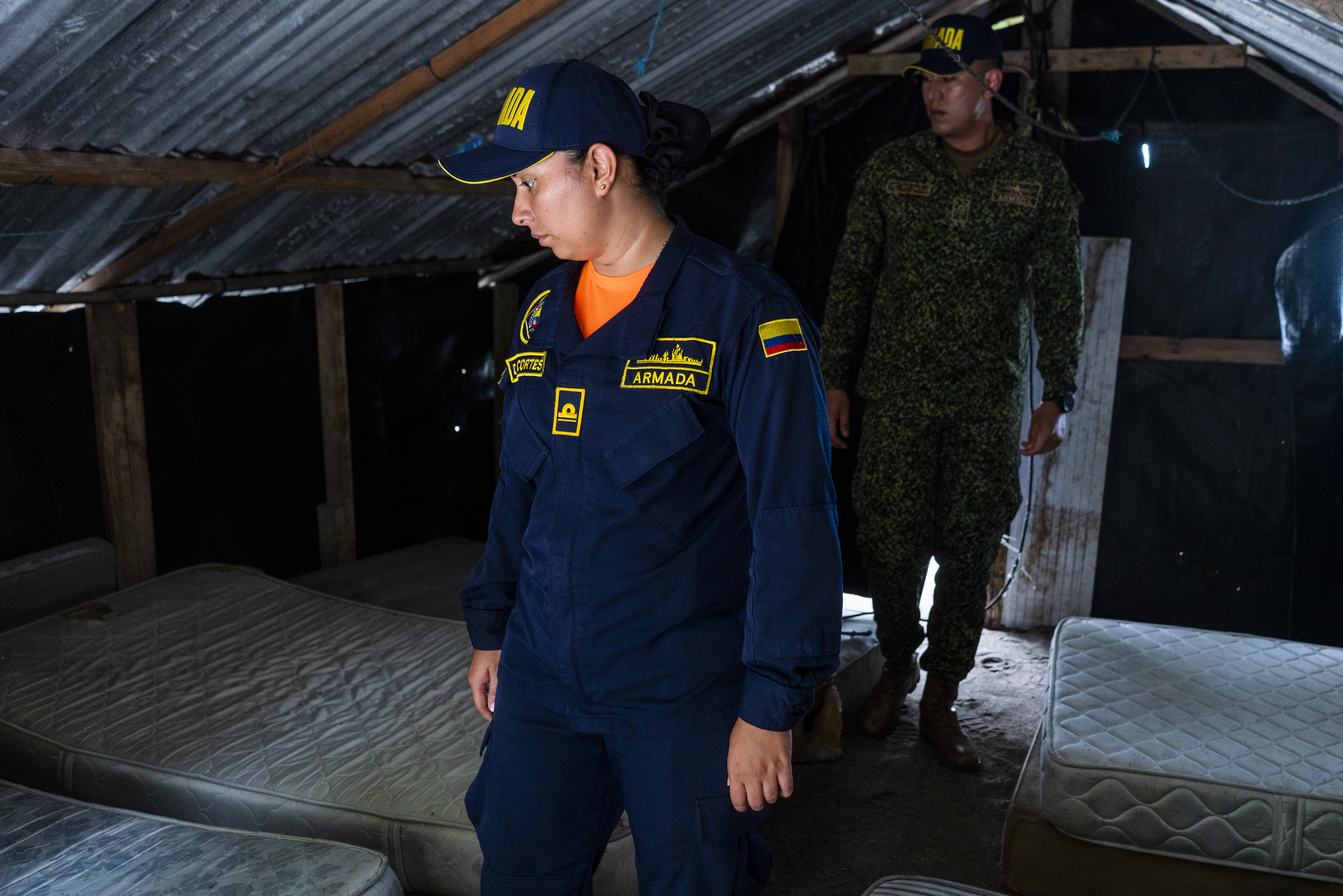 Two naval officers, dressed in black uniforms and caps, walk through a small hut. On either side of them are empty mattresses.