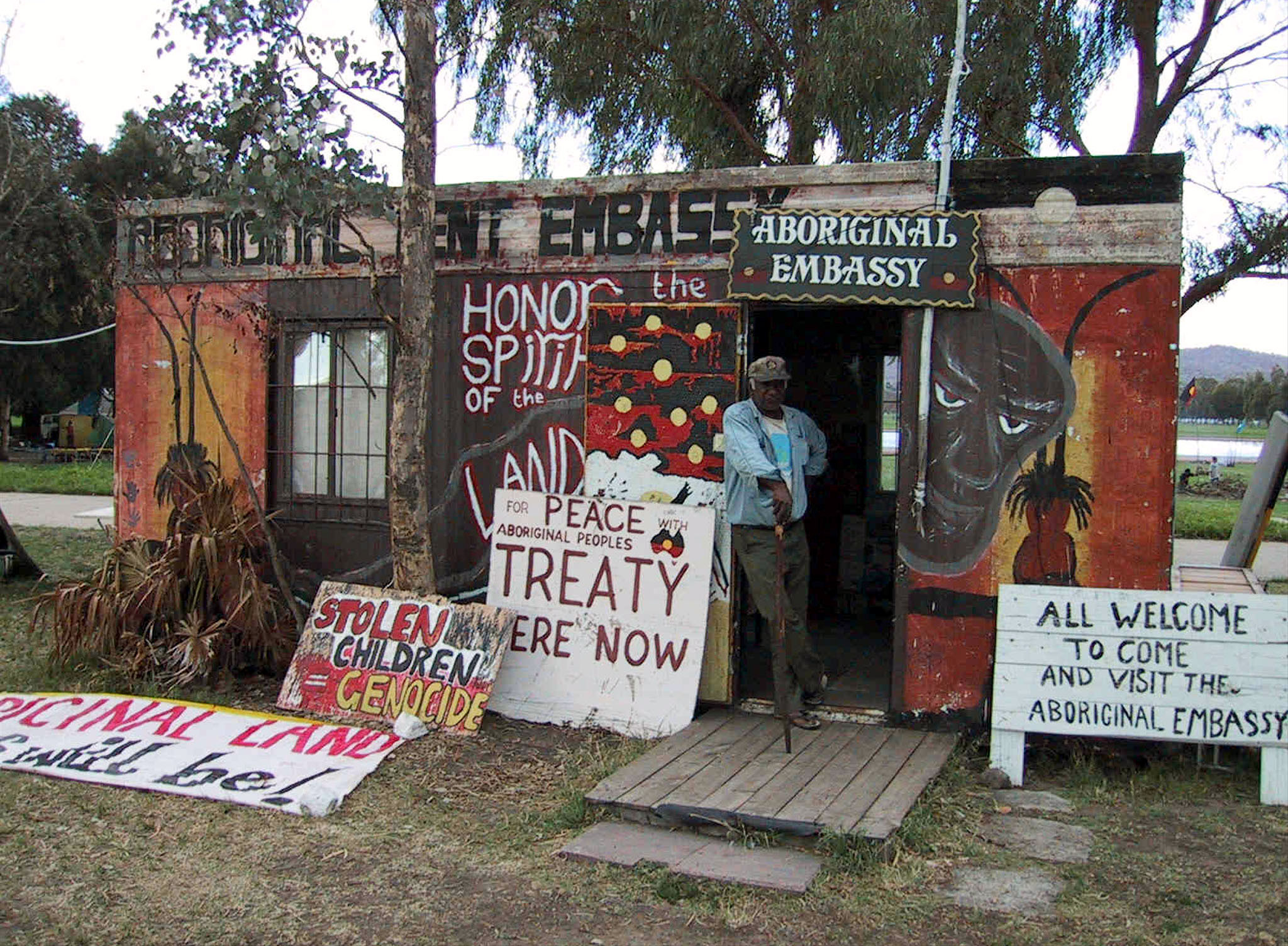 A man leans against the door of a small building with paintings in mostly red and black colours and banners reading Aboriginal Tent Embassy in front of a gum tree and blue sky