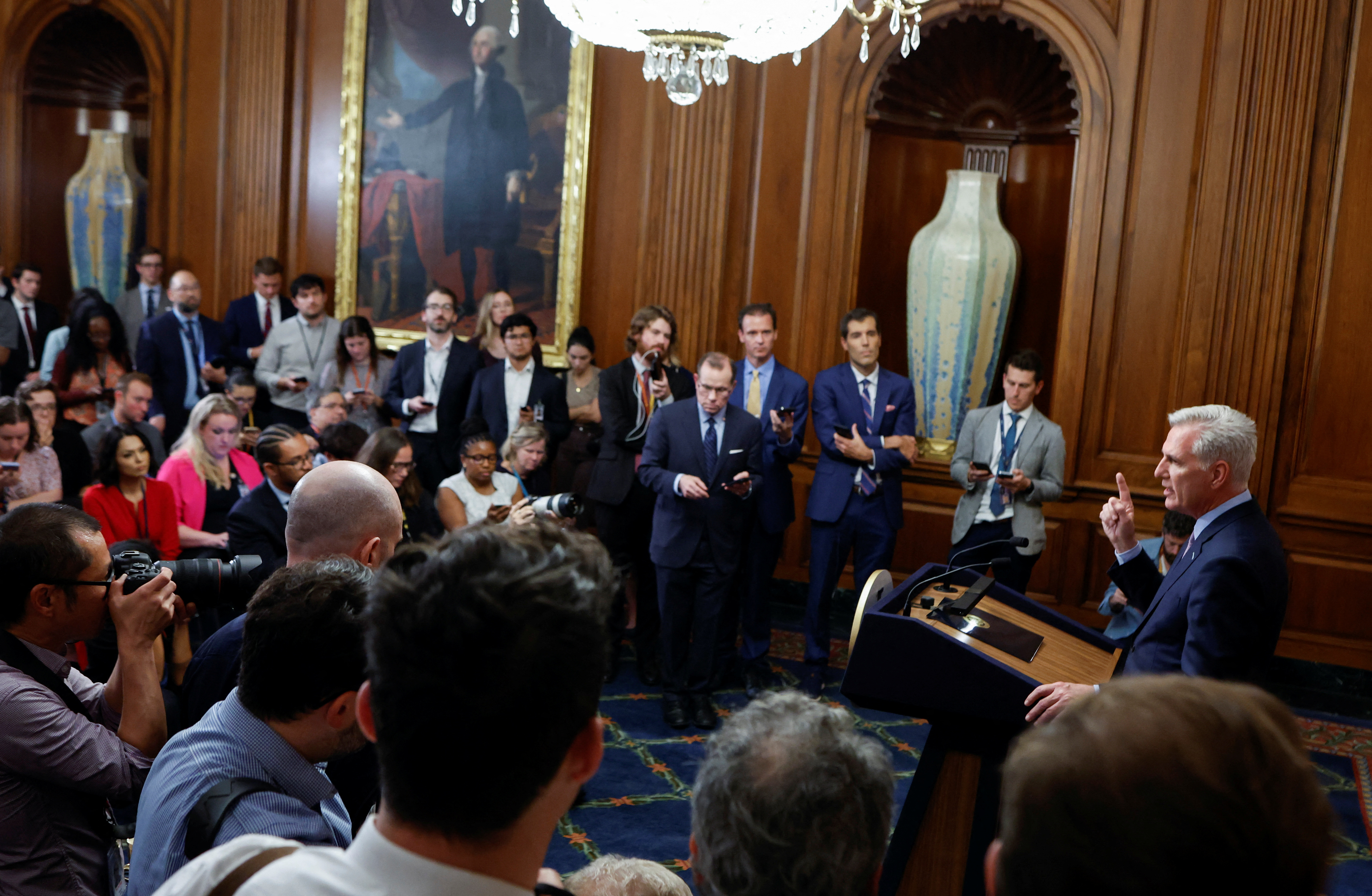 Kevin McCarthy, standing behind a podium, speaks to a ring of reporters in a wood-lined room in the Capitol. He is in mid-gesture, lifting a finger into the air.
