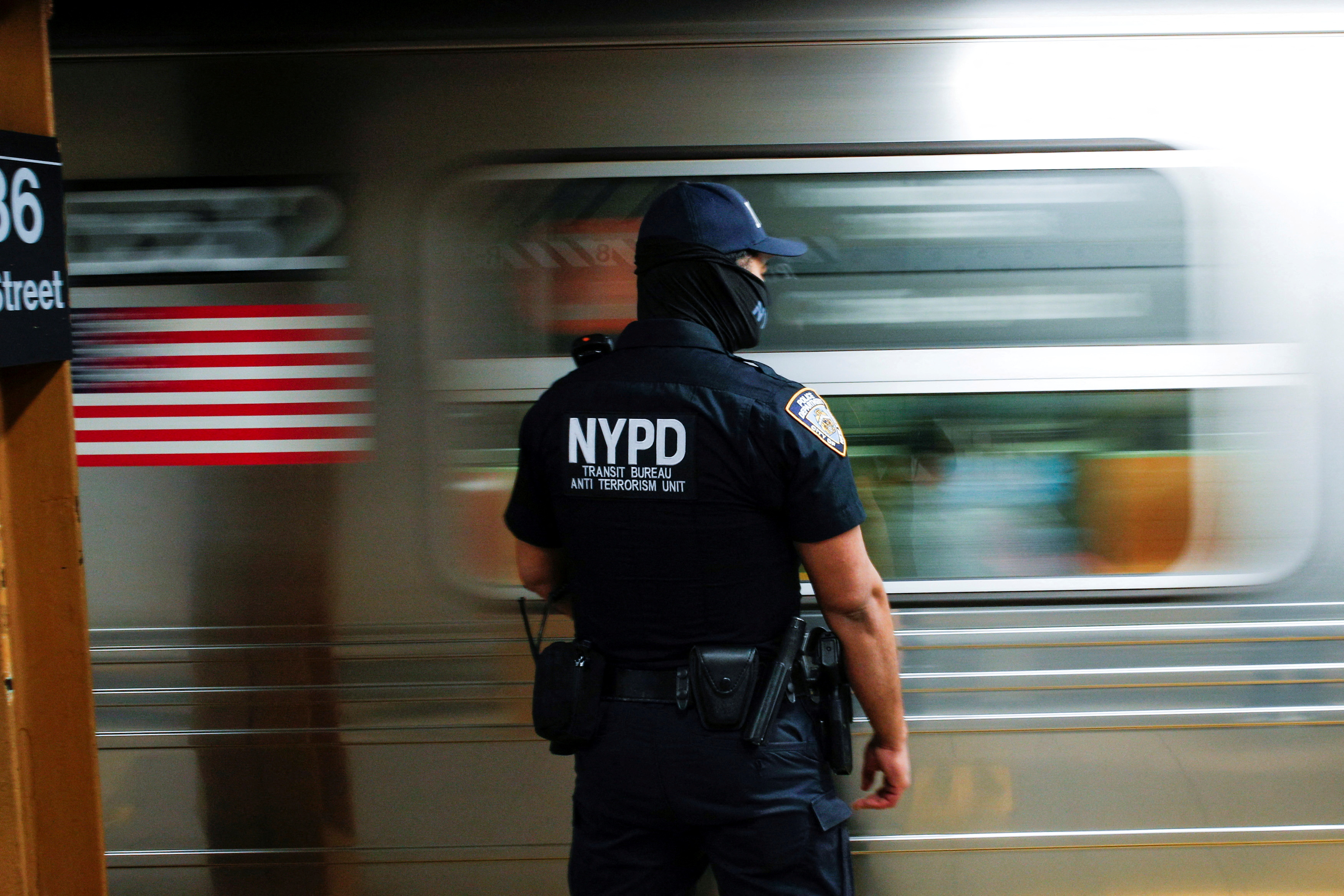 A police officer with "NYPD" written on the back of his uniform stands on a subway platform as a car rushes by.