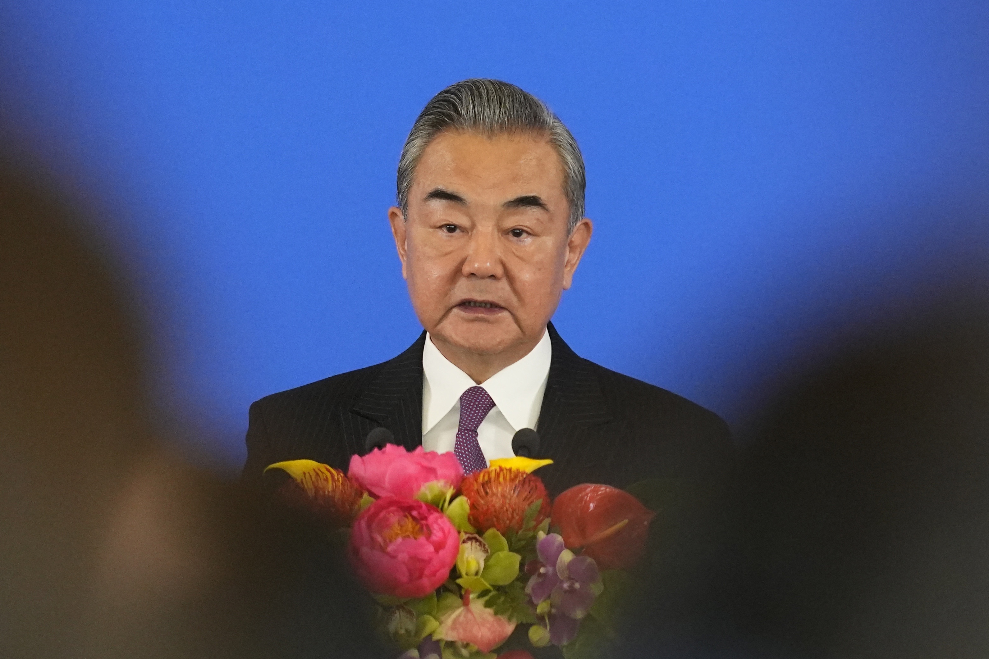 Wang Yi speaking at an event. He has flowers in front of him. The backdrop is blue.