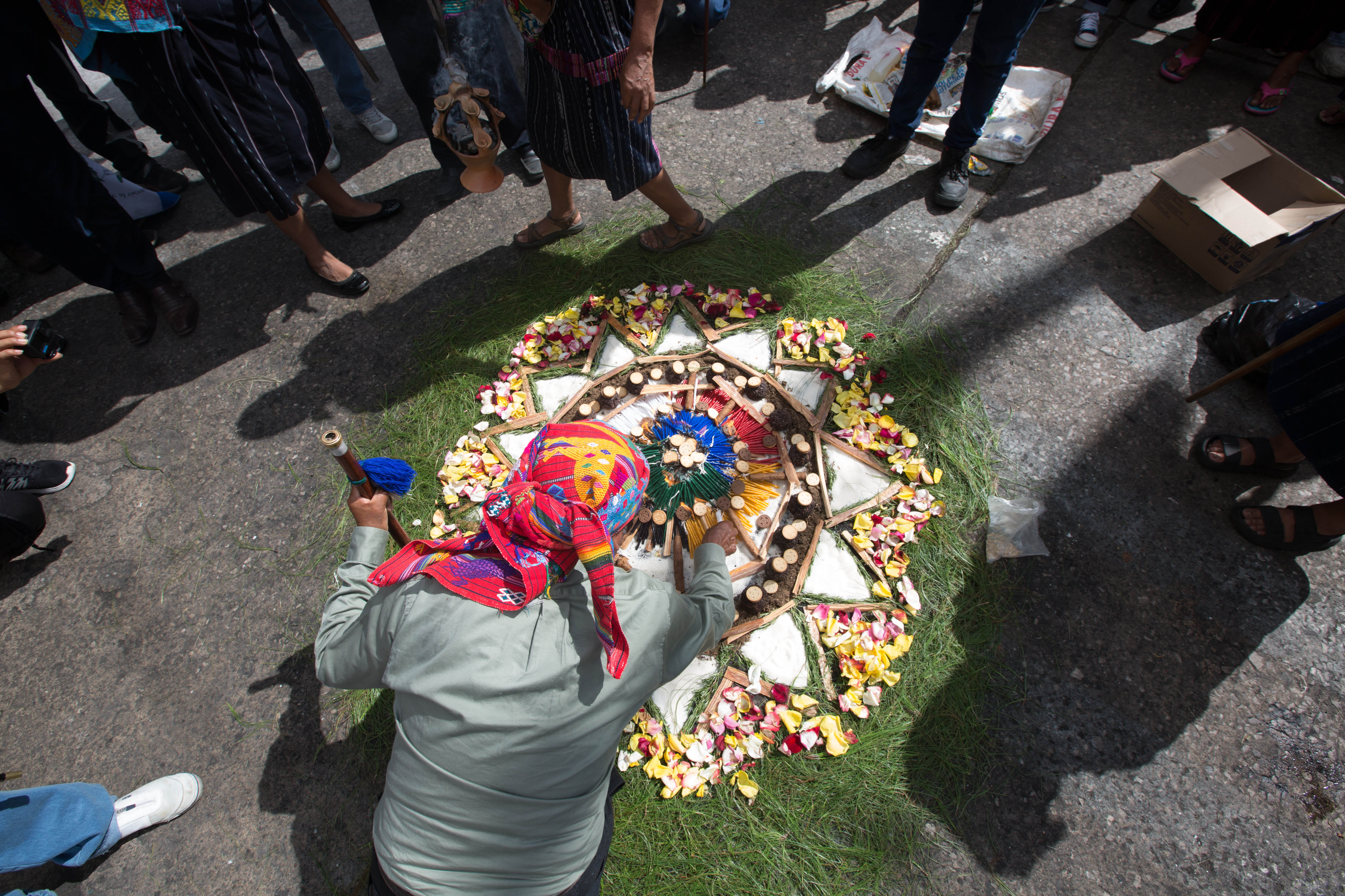 Seen from above, a spiritual leader leans over a circle design on the concrete: The colorful design is green along the edges. Its center has flowers and patterns with white triangles.