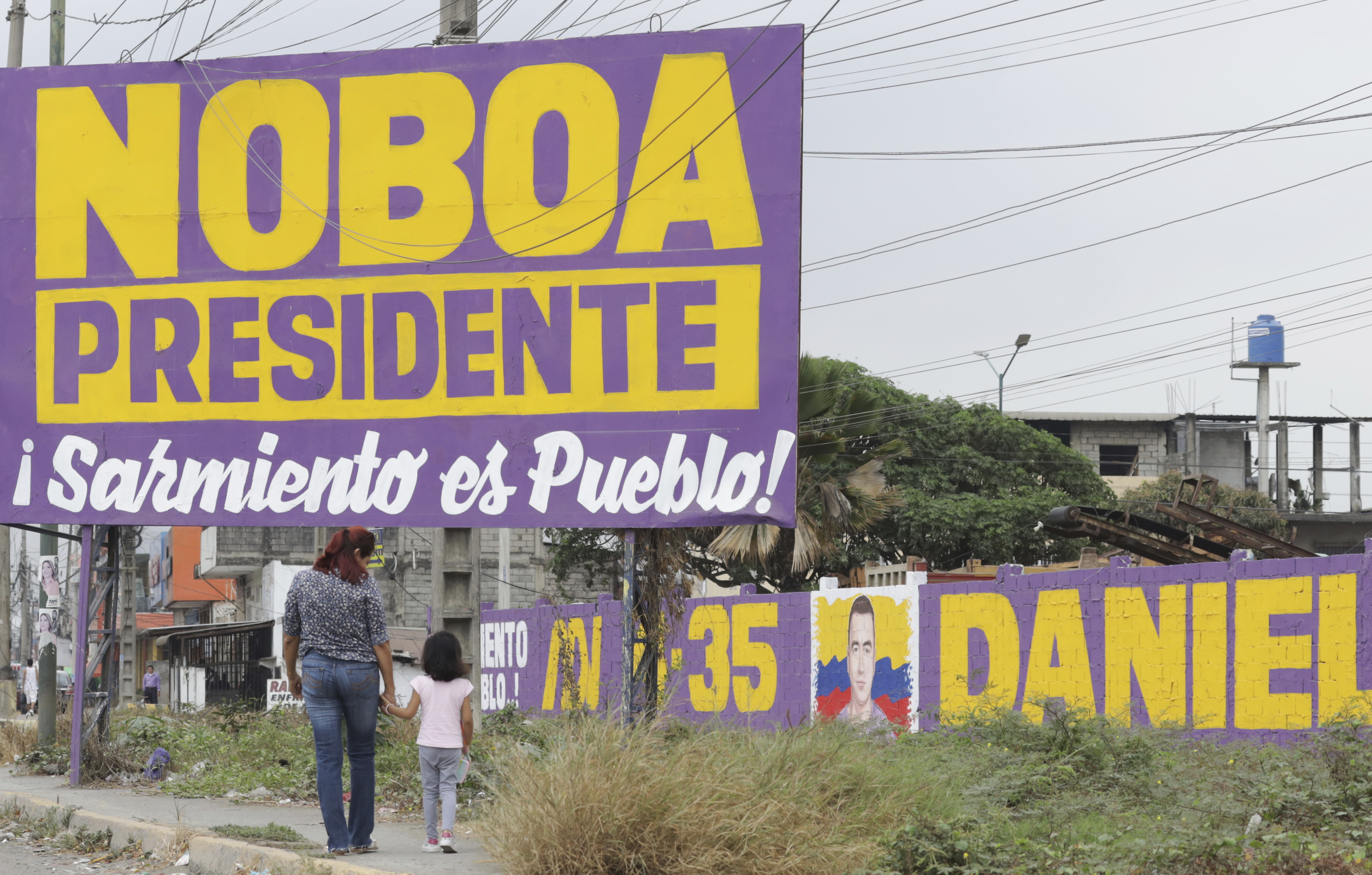 An adult and small child walk along a dirt path next to a billboard and painted wall that read: "Noboa Presidente".