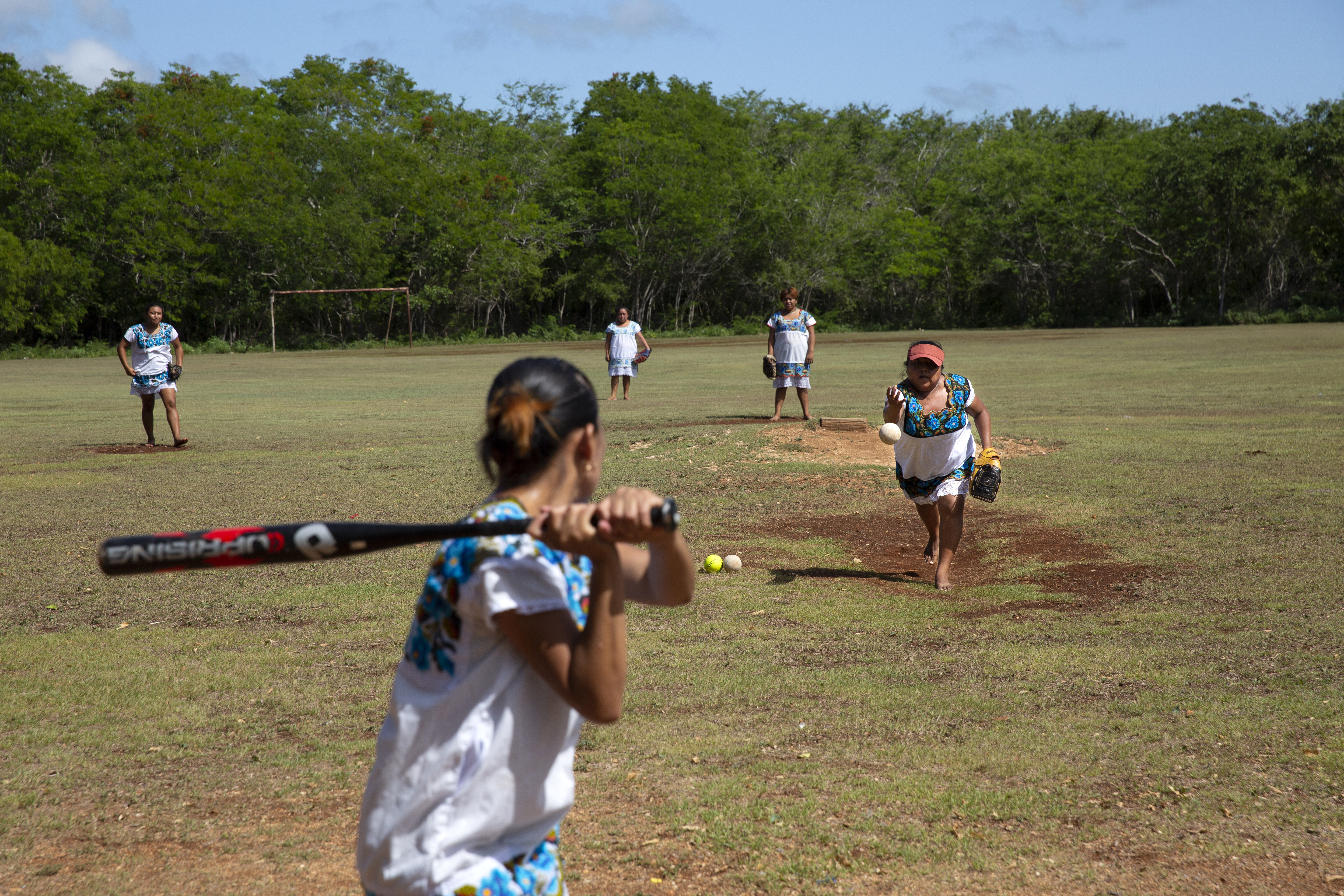 Seen from behind, a young woman waits with ball in hand to swing. A pitcher on a baseball field prepares to throw her the ball.