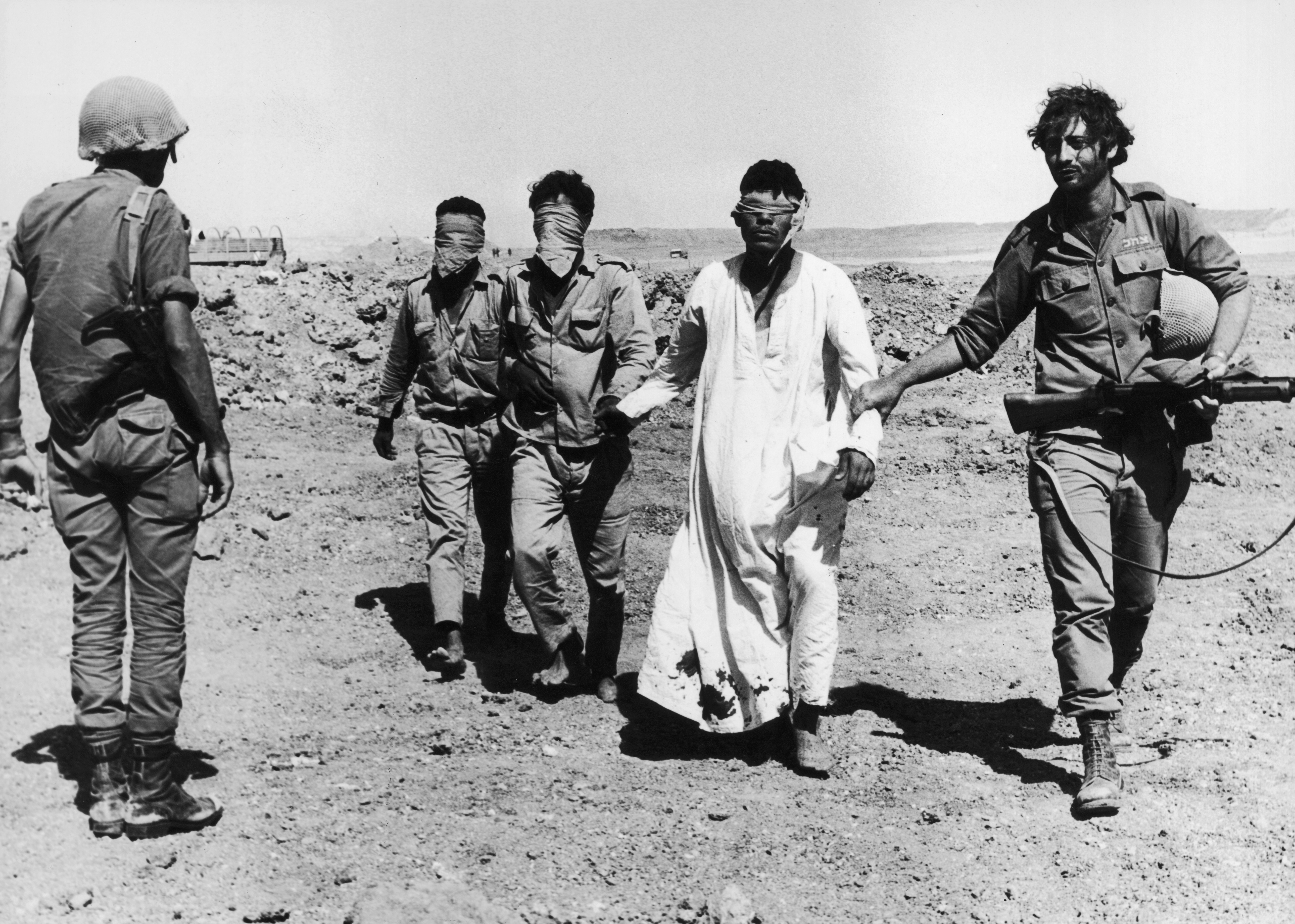 Three barefoot, blindfolded Egyptian POWs are led to the western side of the Suez Canal by an Israeli soldier as another soldier looks on during the Yom Kippur War