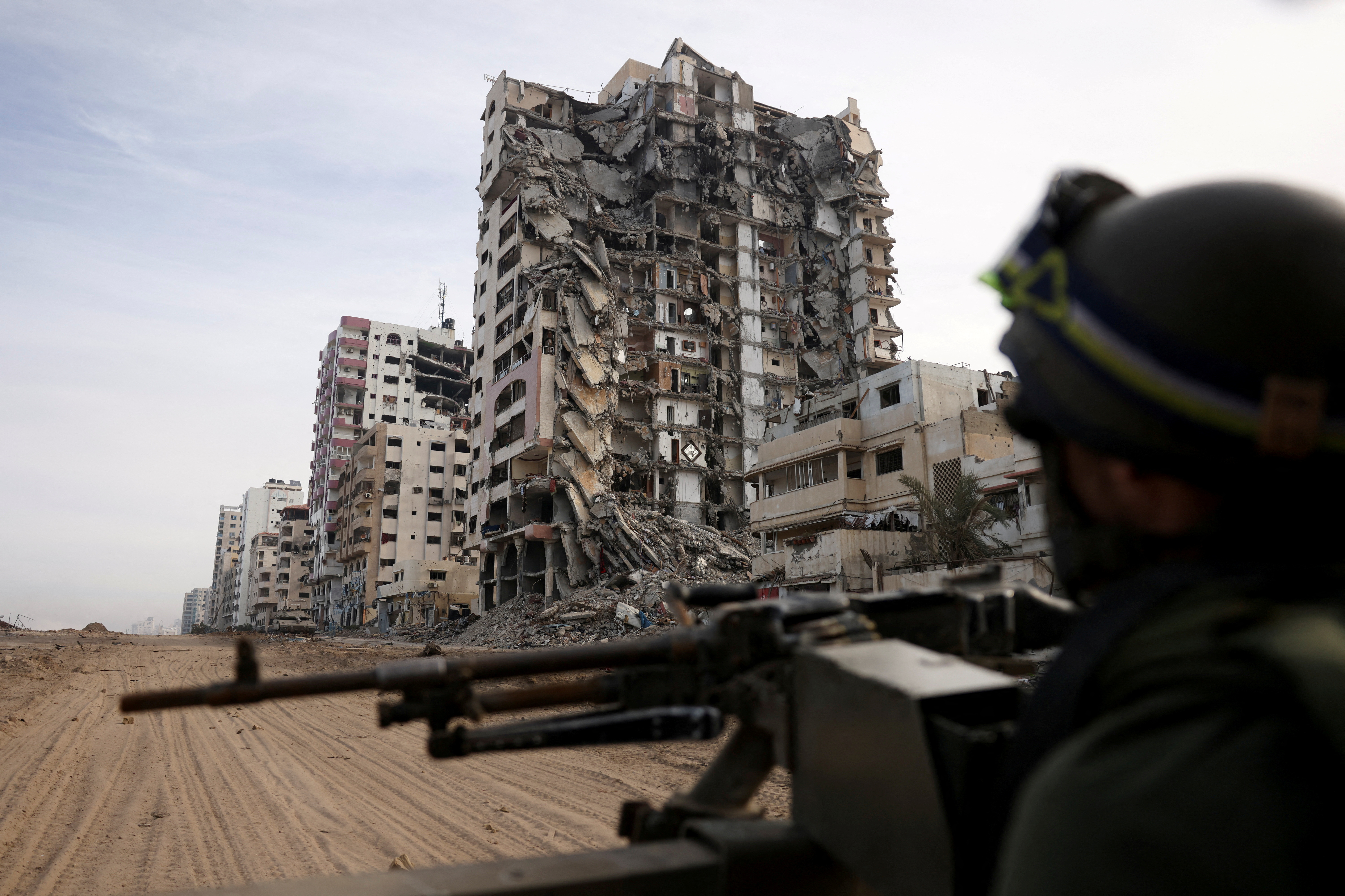 An Israeli soldier sits on a Humvee in northern Gaza. There are collapsed apartment blocks on one side. 