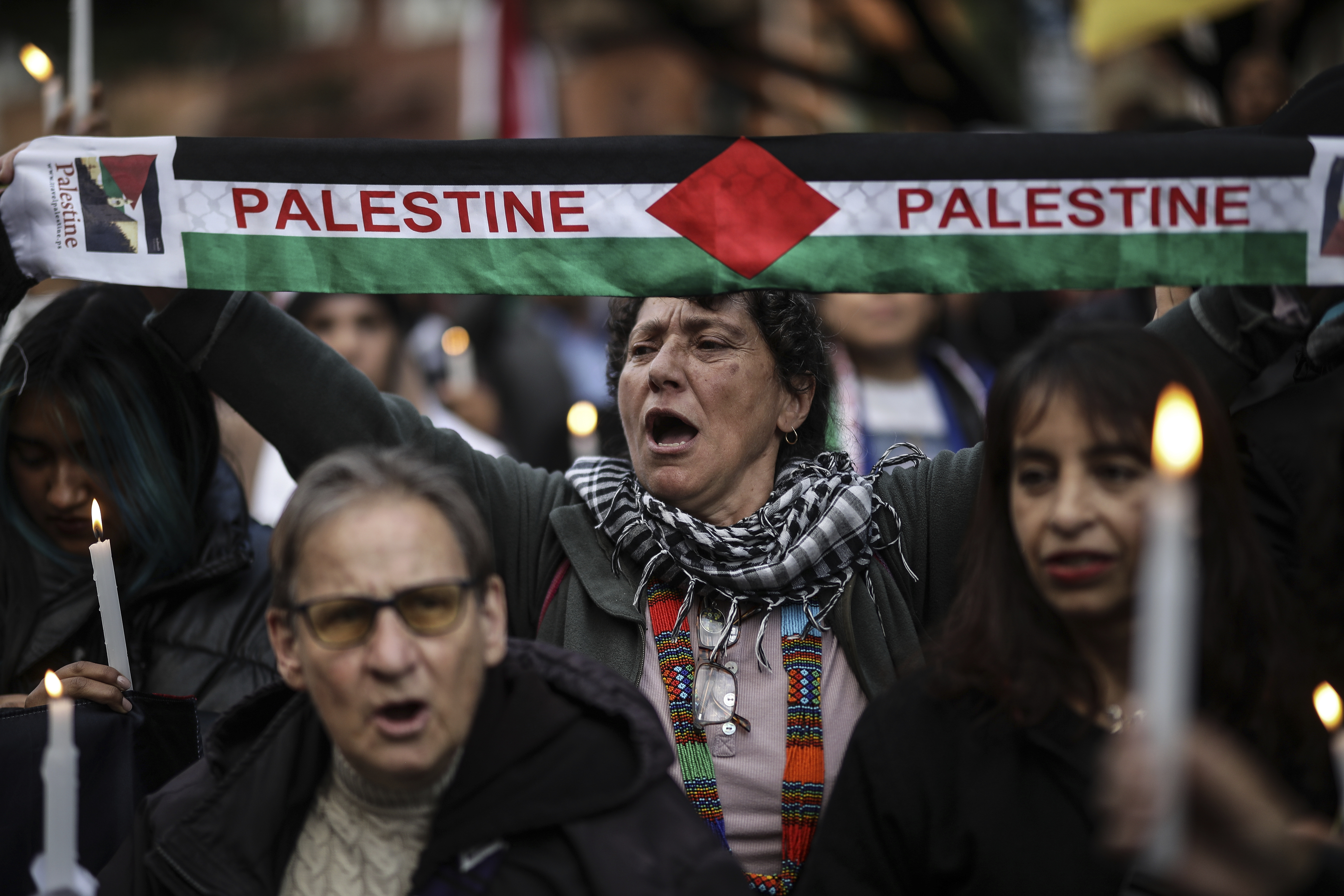 A woman holds up a scarf in the colors of the Palestinian flag, with the word "Palestine" written in red. Around her, people carry candles.