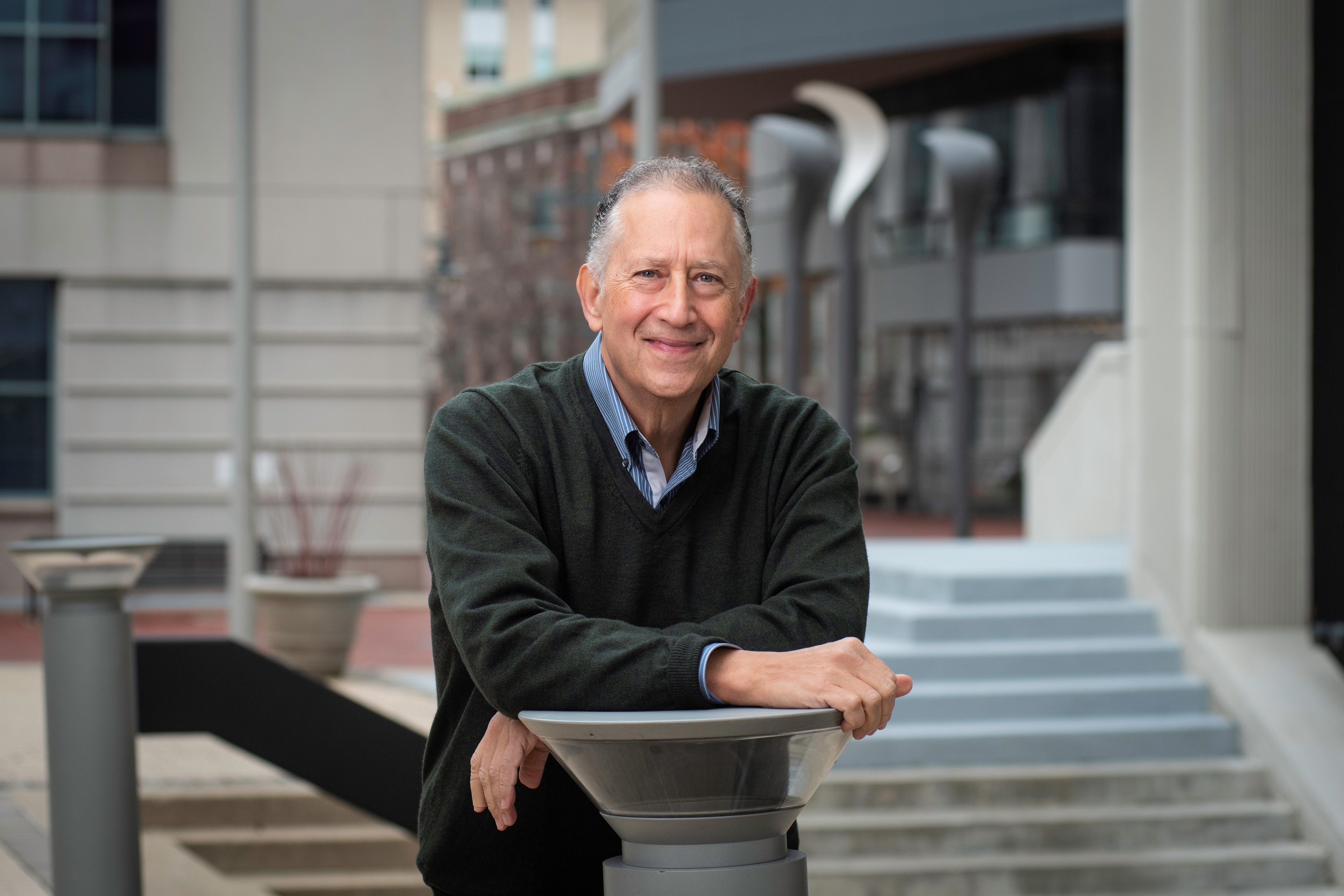 A man in a grey pullover sweater, collared shirt and tie leans against a railing outside of an academic institution.