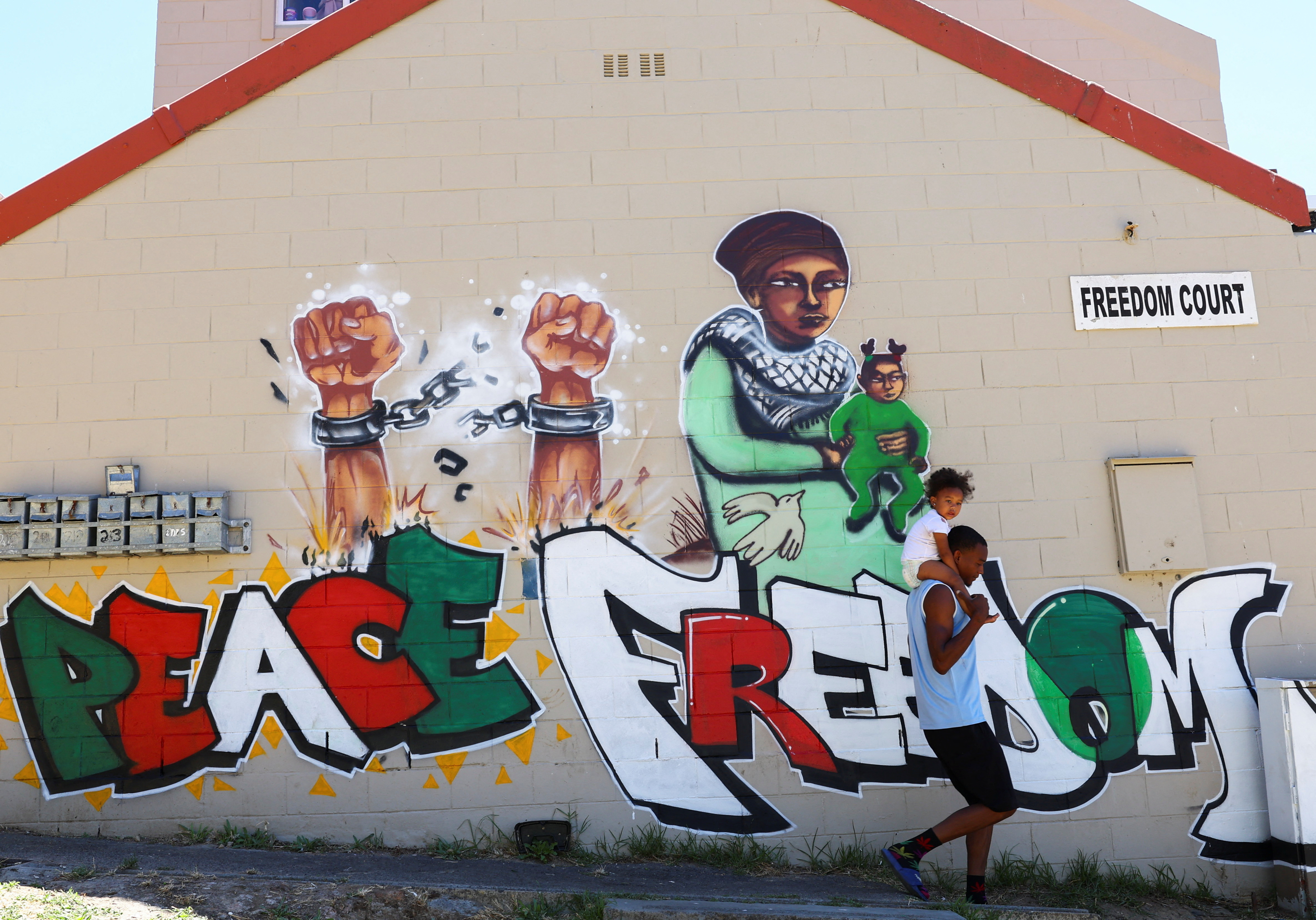 A man carrying a child walks past a mural in Bo-Kaap symbolising freedom and peace in support of Palestinians in Gaza