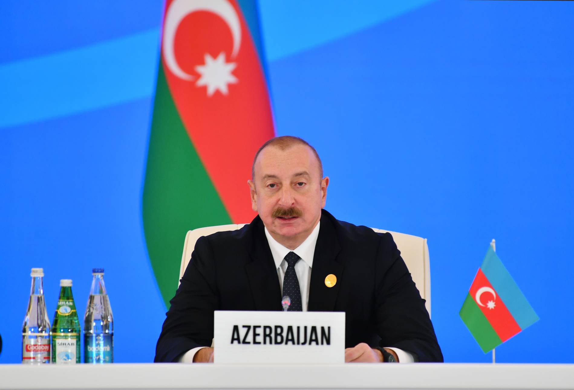 A photo of Ilham Aliyev sitting at a table with a sign that says "Azerbaijan" in front of him with three bottles of water next to him as well as a small Azerbaijan flag next to him and a bigger Azerbaijan flag behind him.