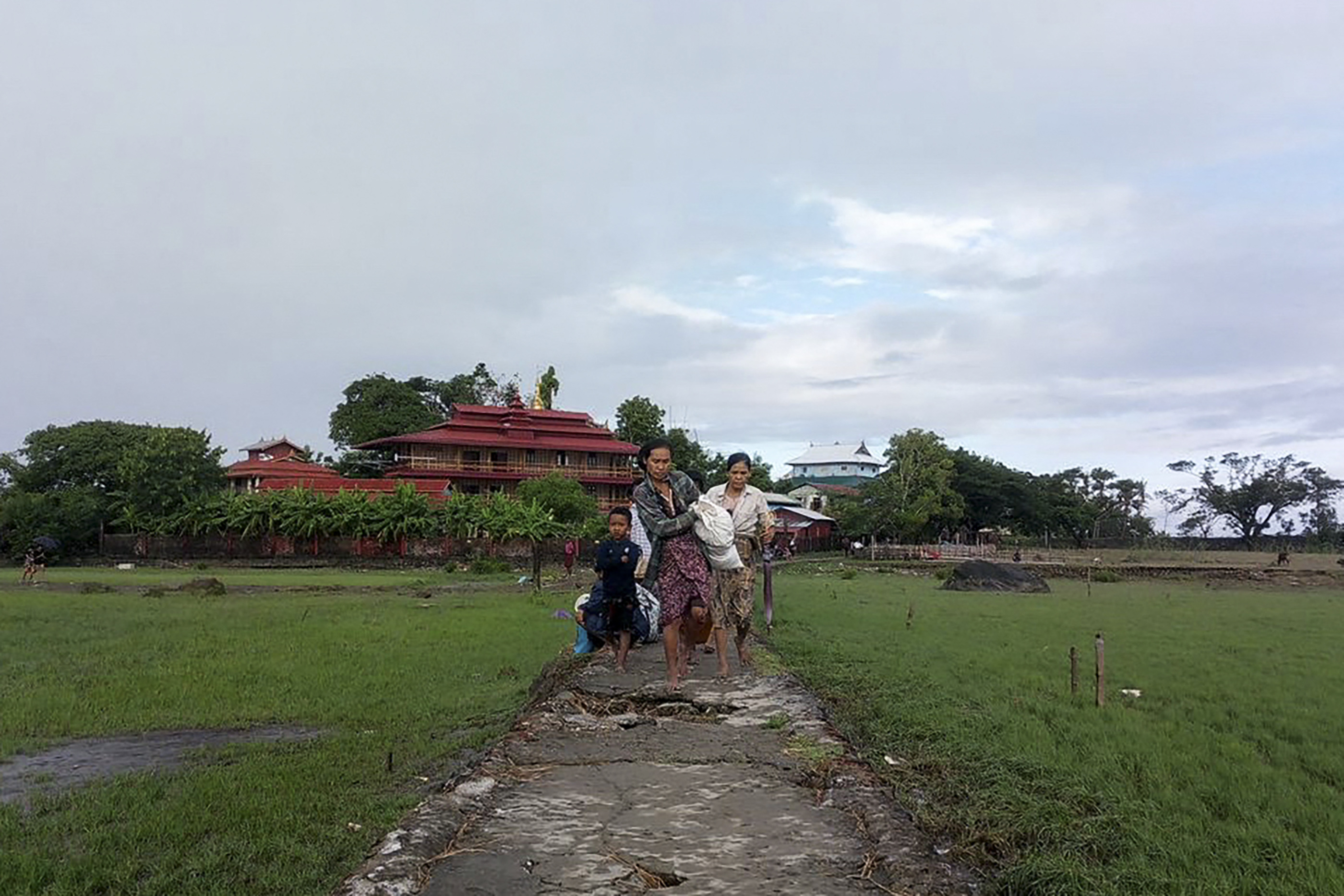 People running from a village in Rakhine state. They are carrying sacks of belongings and are on a pathway through fields. There are buildings behind them.