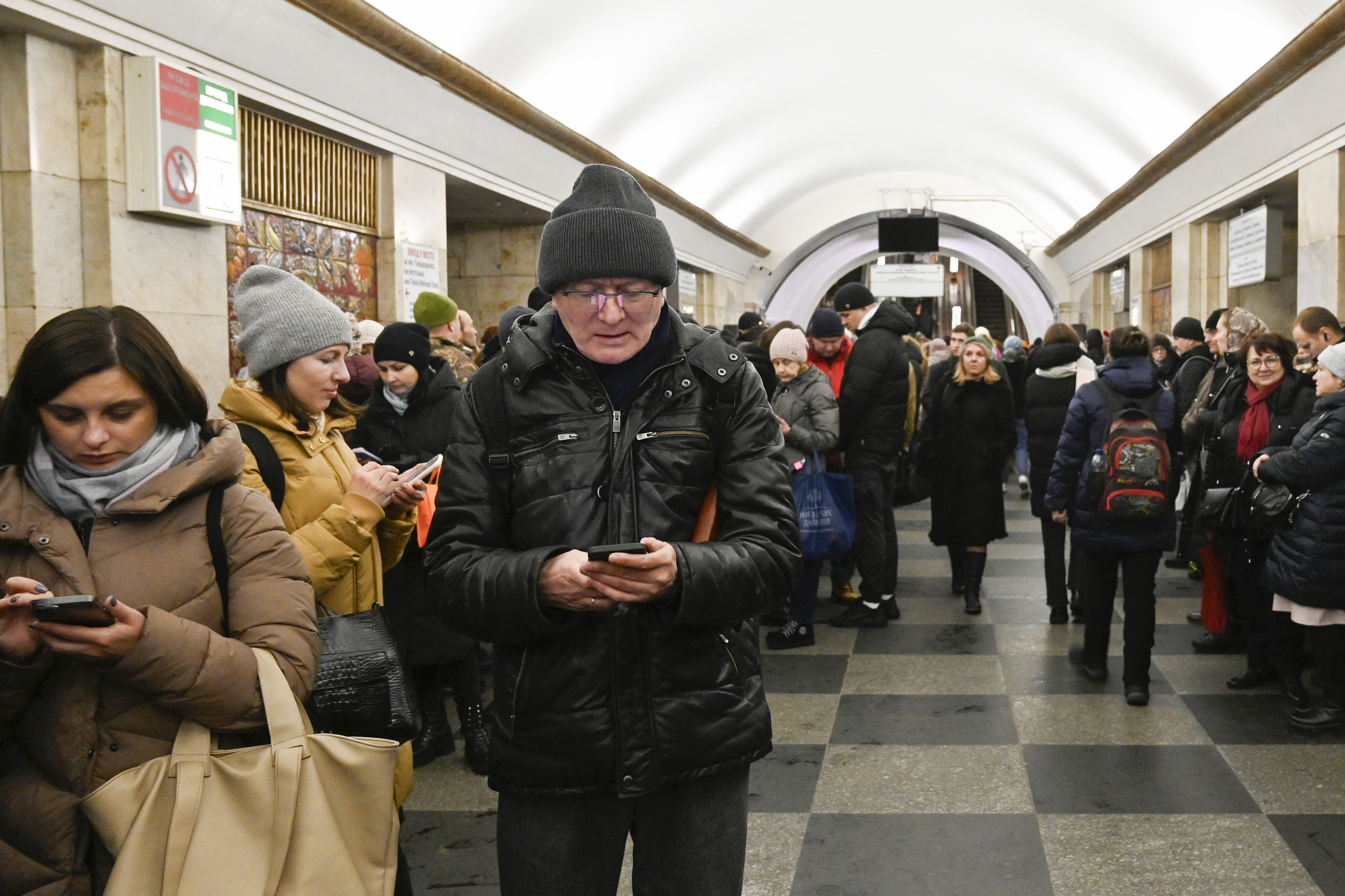 People taking shelter in the Kyiv metro during an air rad. They are dressed in winter clothes. Some are looking at that phones.
