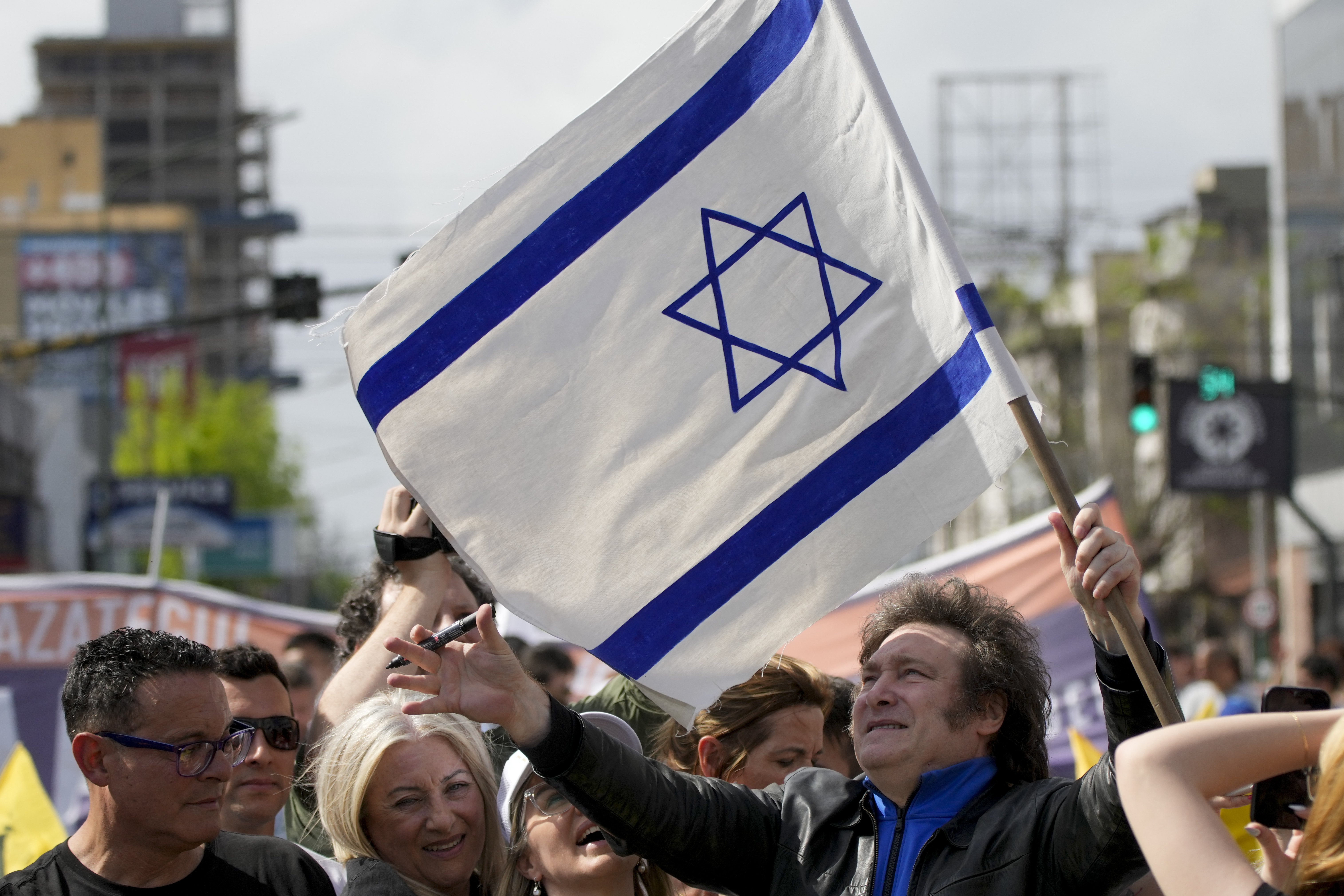 Javier Milei, standing outdoors amid a crowd, holds up an Israeli flag.