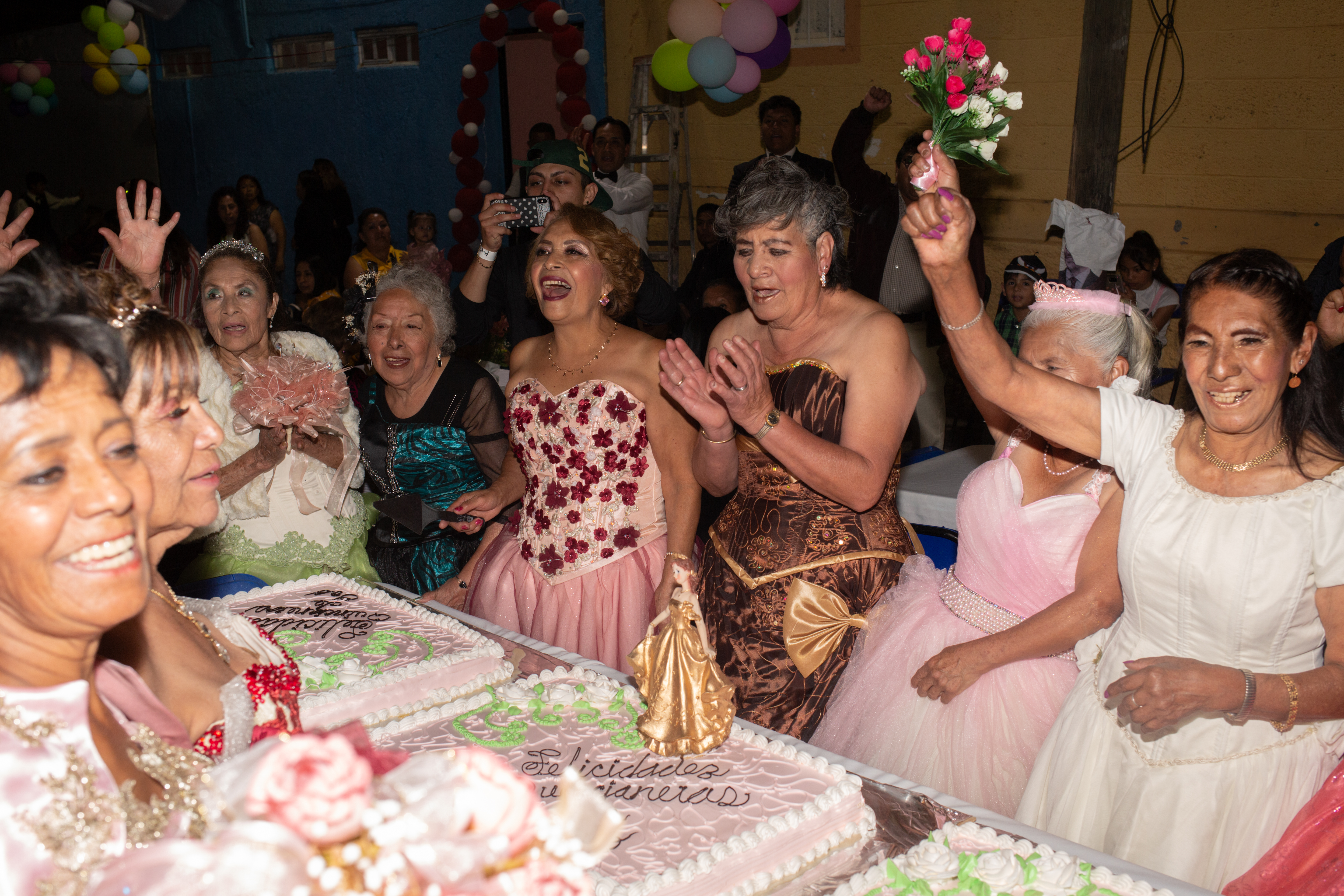 Women in opulent ballgowns — some bronze with gold trim, others pink with red flowers — circle around a table covered with three sheet cakes, covered in icing and decorations. One woman raises a bouquet of flowers into the air.