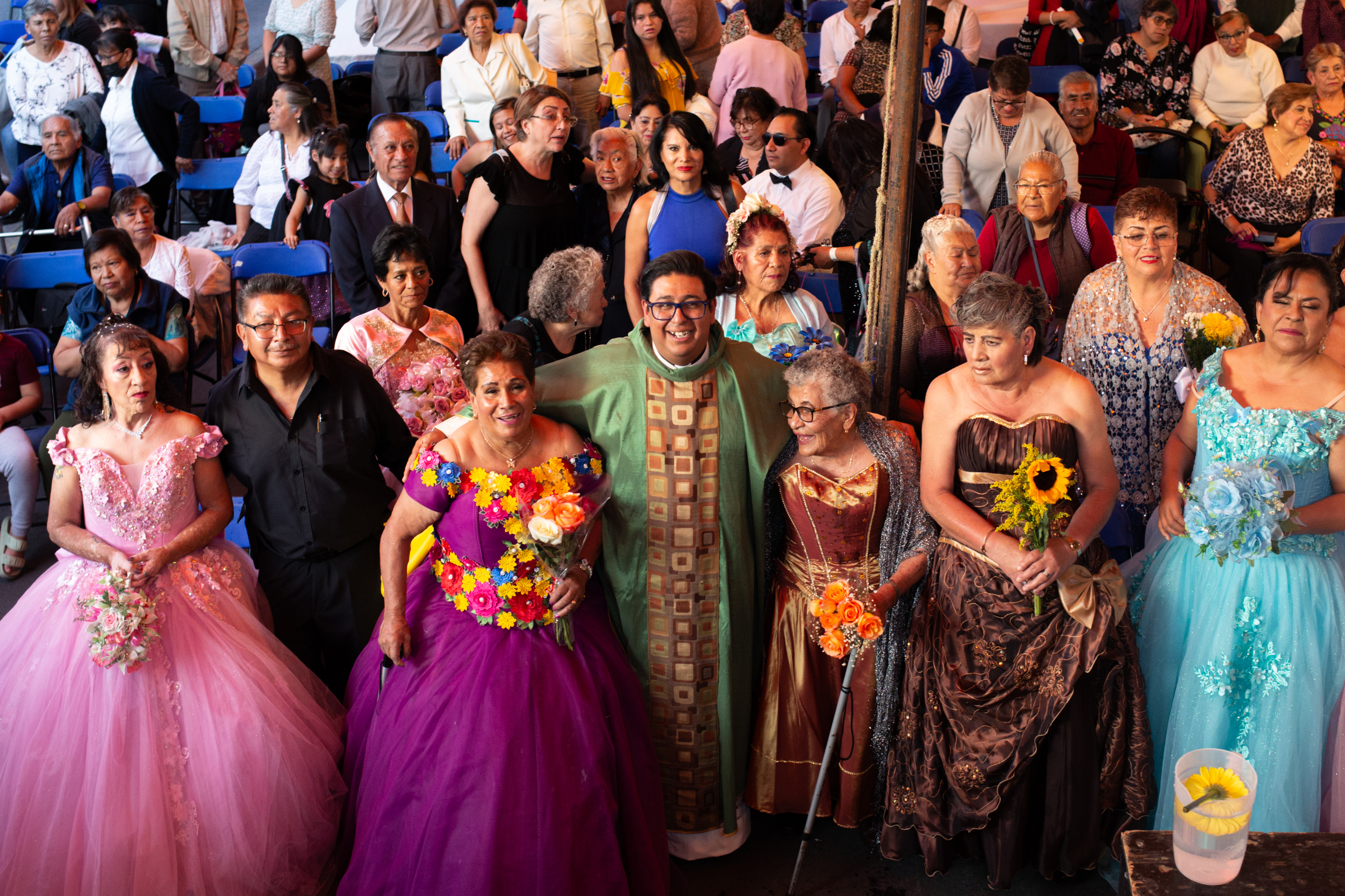 A priest in a green frock with gold trim stands amid a sea of women dressed in beautiful, multi-coloured ballgowns.