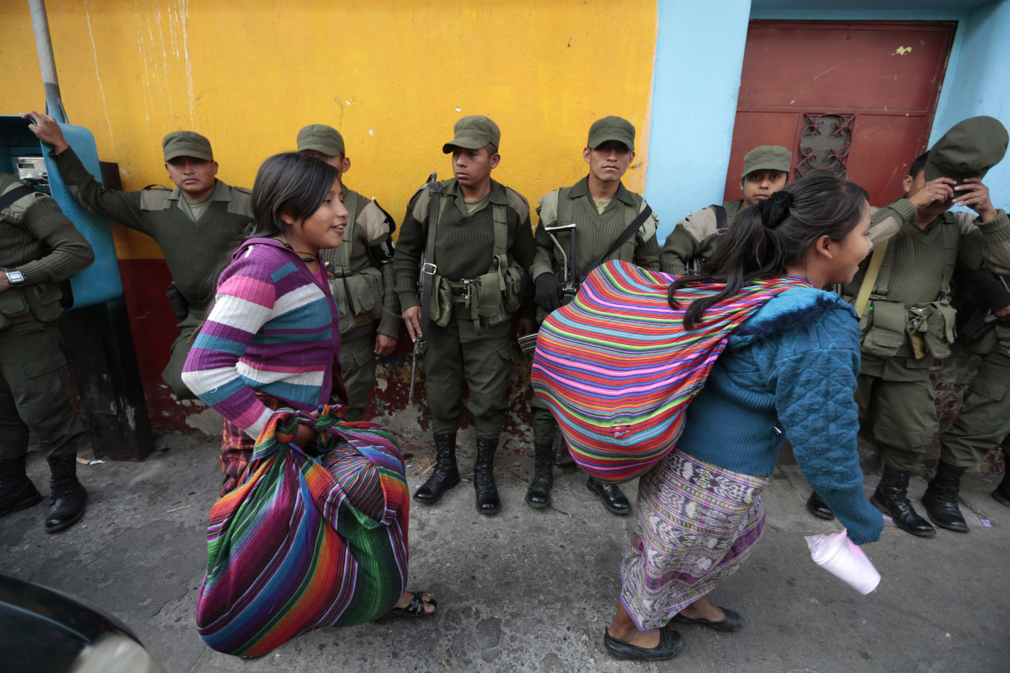 Women in traditional textiles — with rainbow-colored stripes — walk in front of a line of military members on a Guatemalan street.