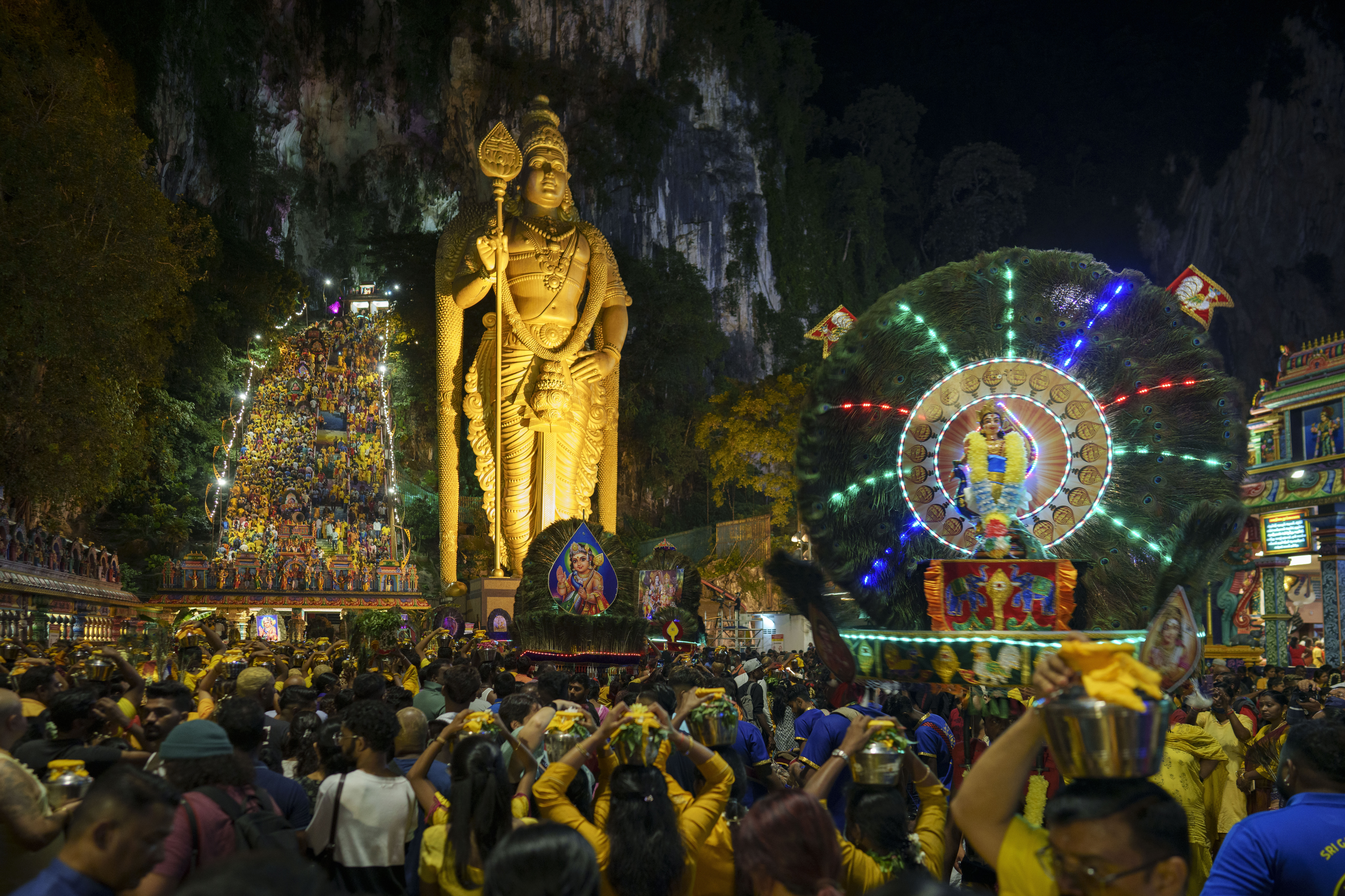 A view of the Batu Caves temple. There are steep steps up to the cave on one side crowded with people. On the right is a towering golden statue of Lord Murugan, In front there are huge crowds of people walking towards the temple with milk pots on their heads