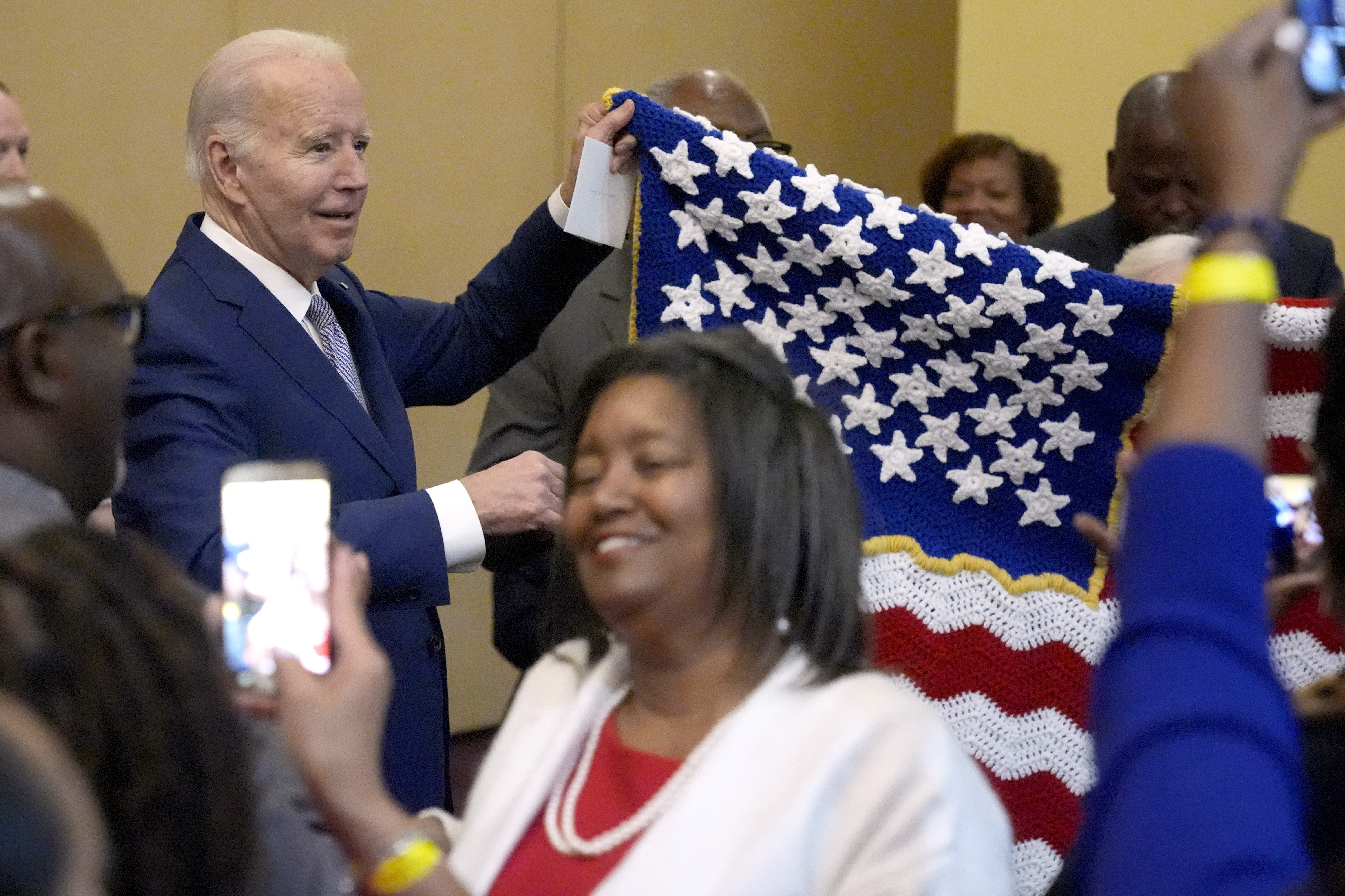 Joe Biden, dressed in a dark suit, holds up a crocheted US flag, while a woman poses for a cellphone photo.