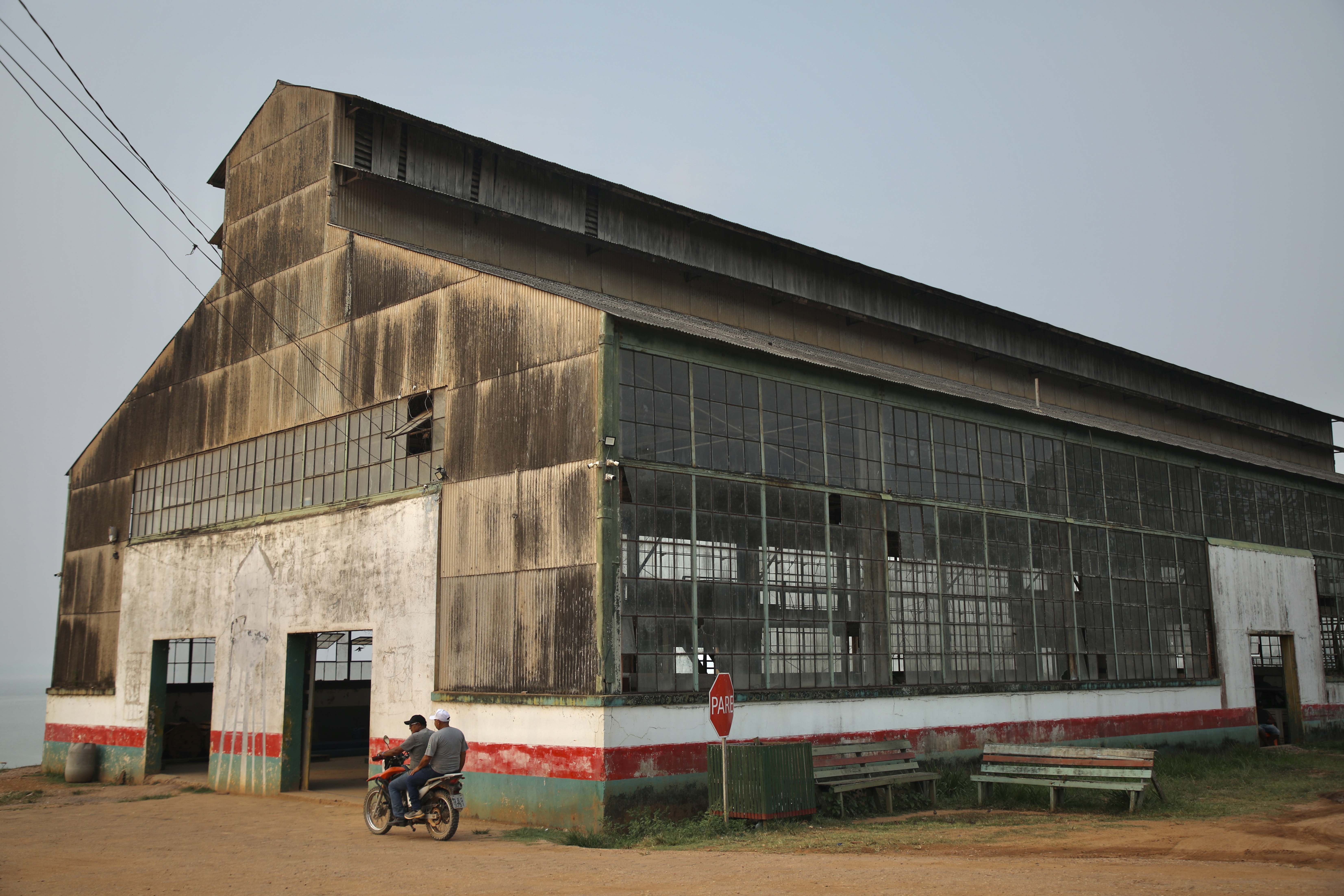 A large warehouse with broken windows sits on a dusty patch of cleared earth.
