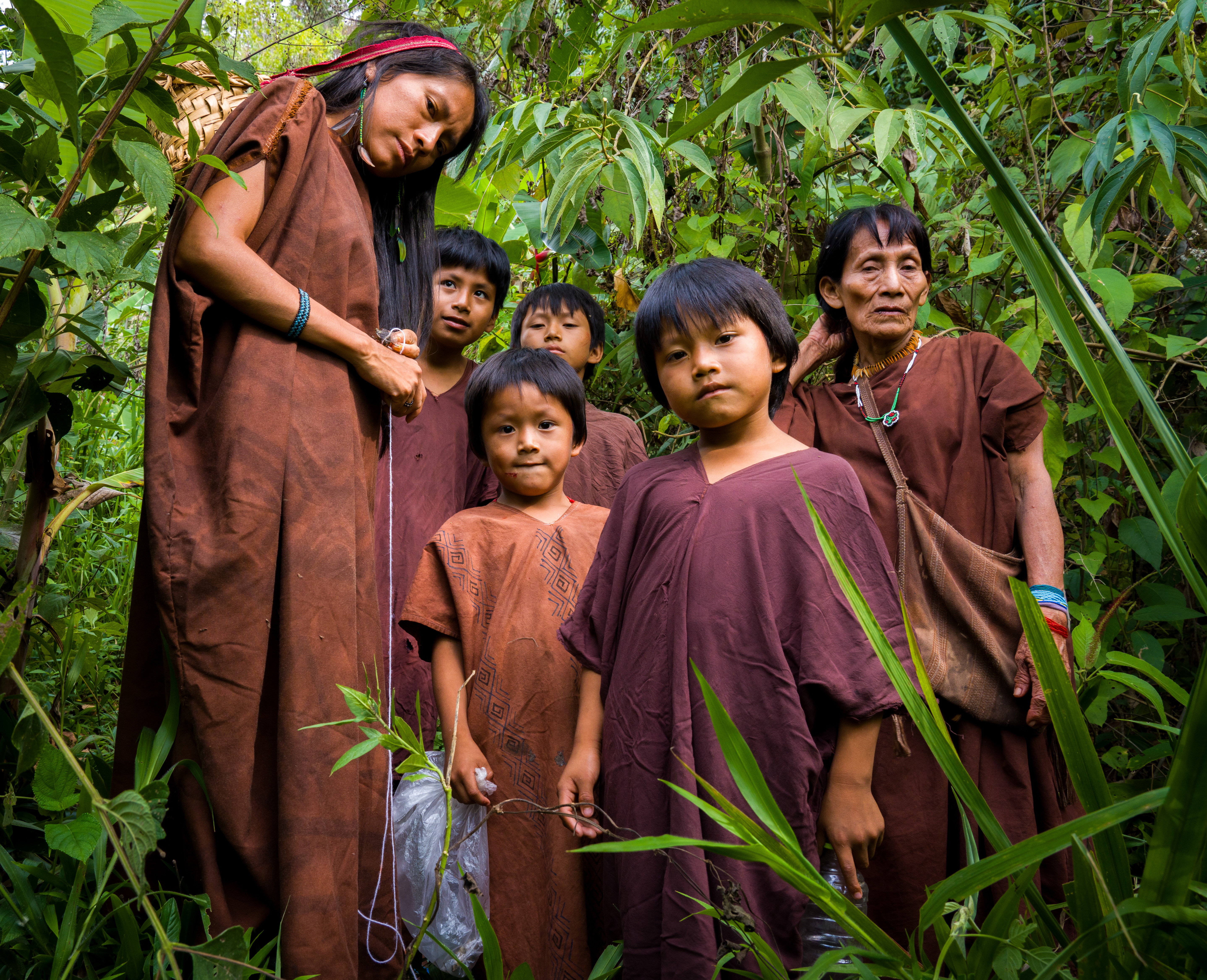 A family from the Ashaninka village of San Miguel Centro Marankiari stand in the greenery of the Amazon rainforest. They include two adult women — one older than the other — and four small children.