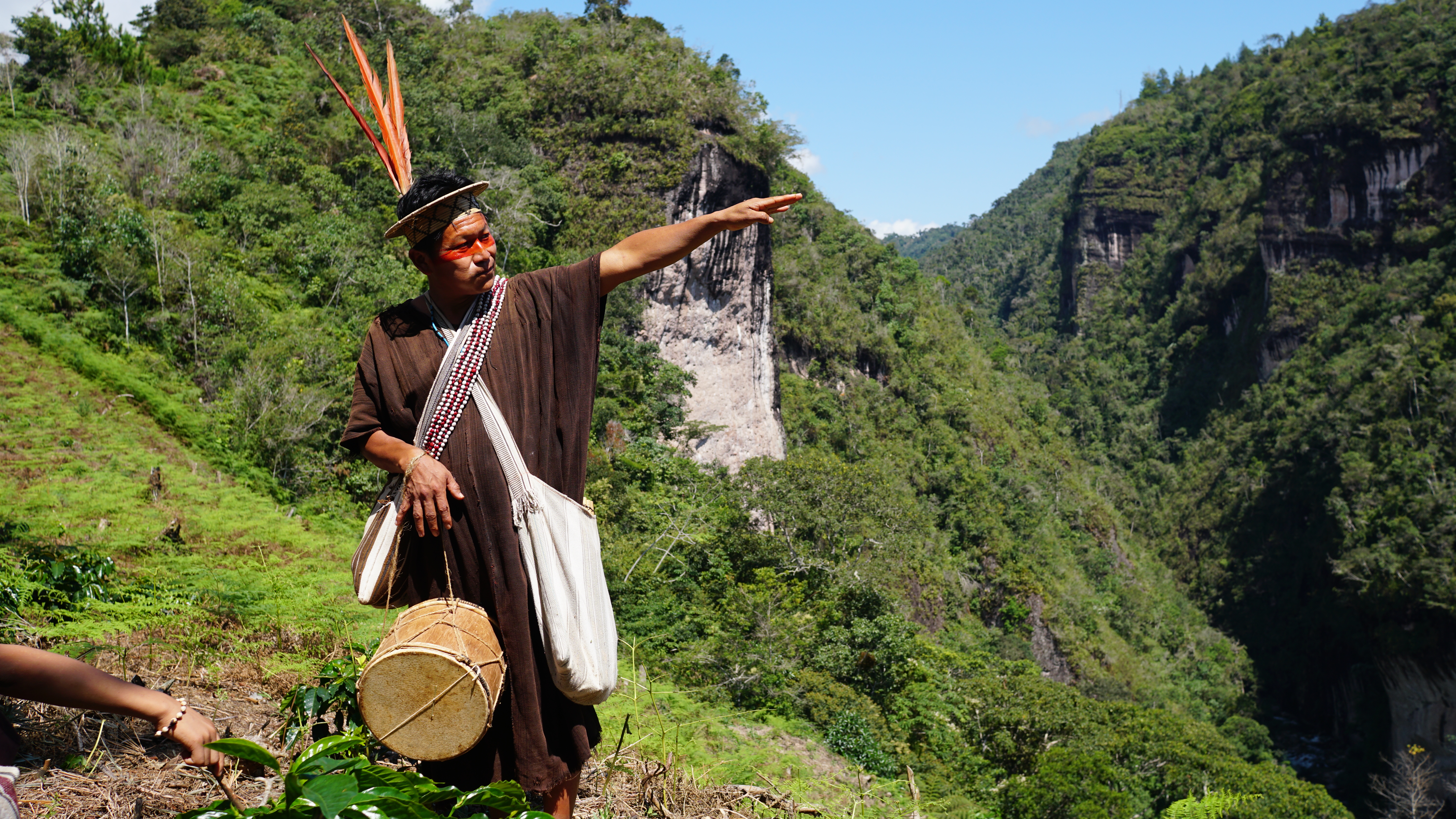 An Indigenous Ashaninka man, dressed in a straw hat topped with three upright red macaw feathers, points over the forested mountains of Peru's Amazon.