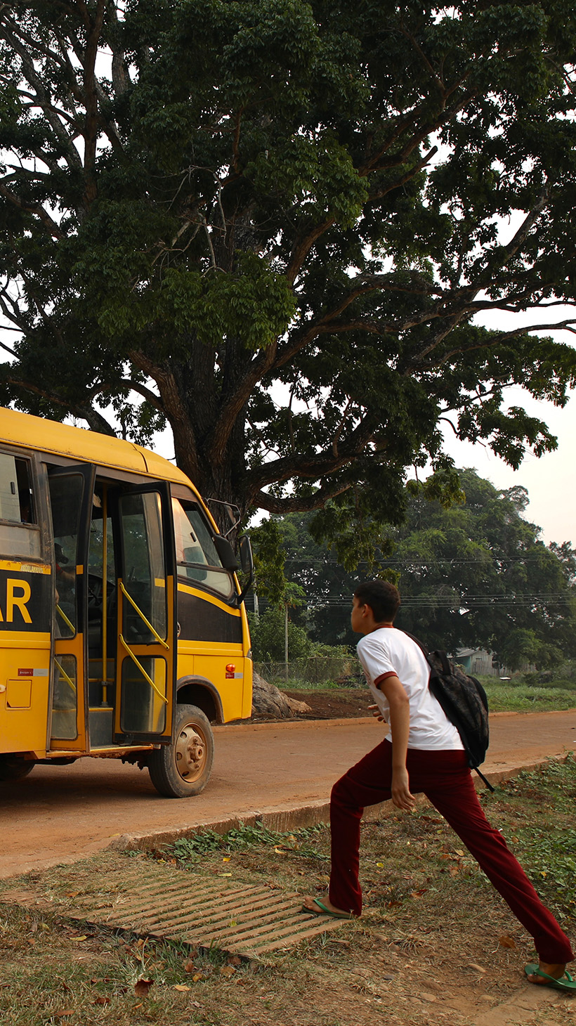 A young student takes wide strides to reach an orange school bus waiting for him on a dirt road.