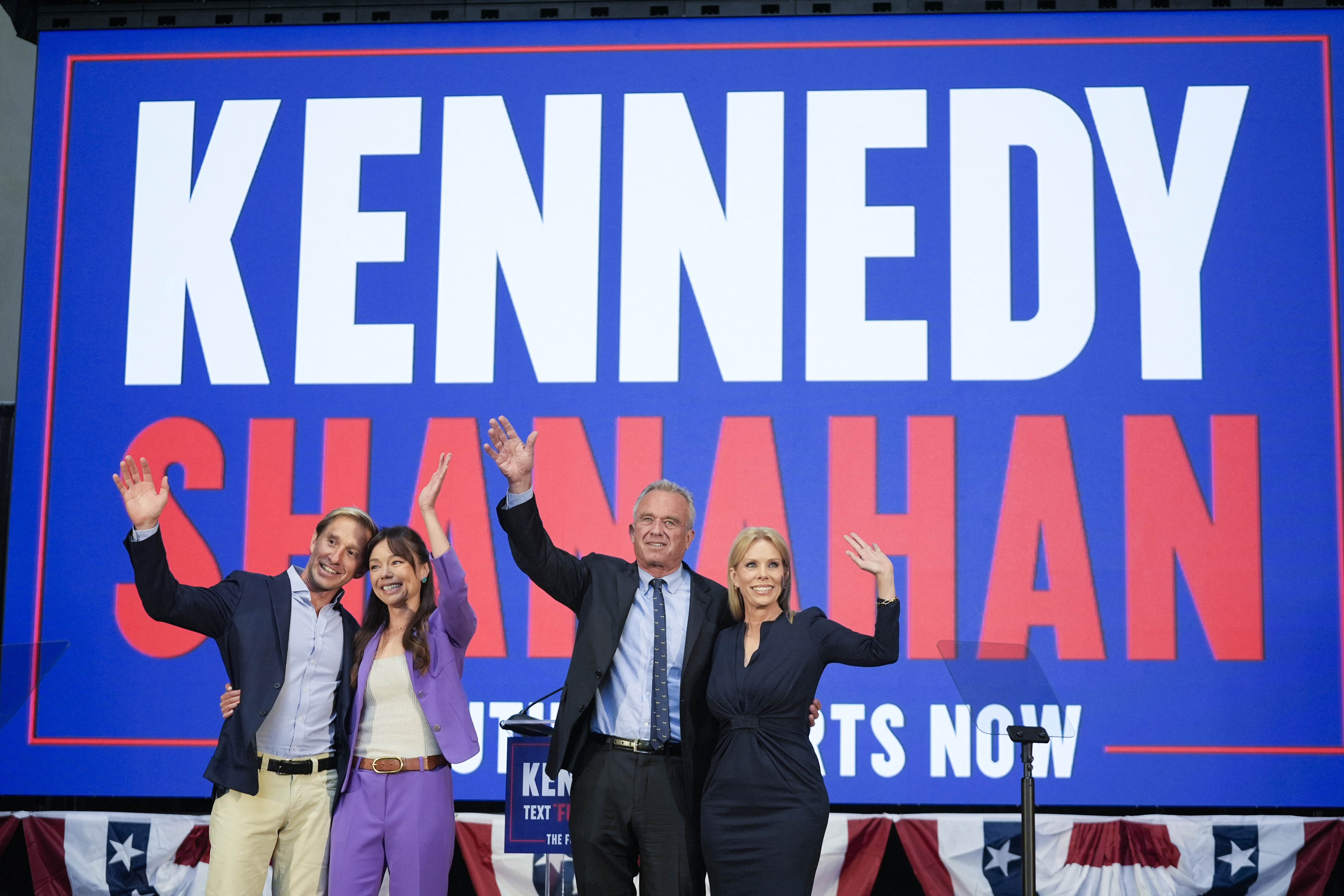 Nicole Shanahan and Robert F Kennedy hug their spouses in front of a screen that displays their names.