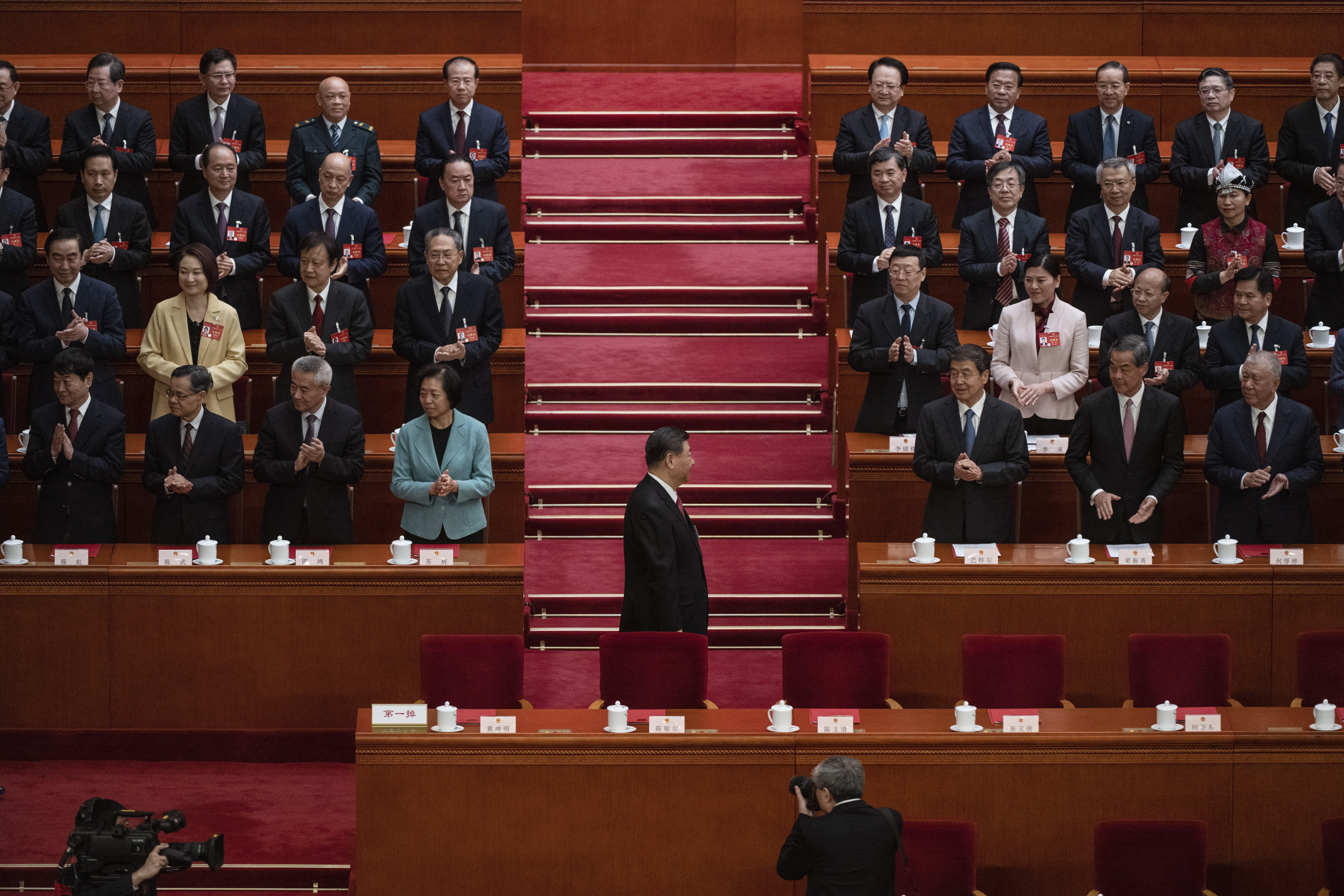 a man in a suit walks past people in dark clothes standing in neat rows in a hall with red carpet