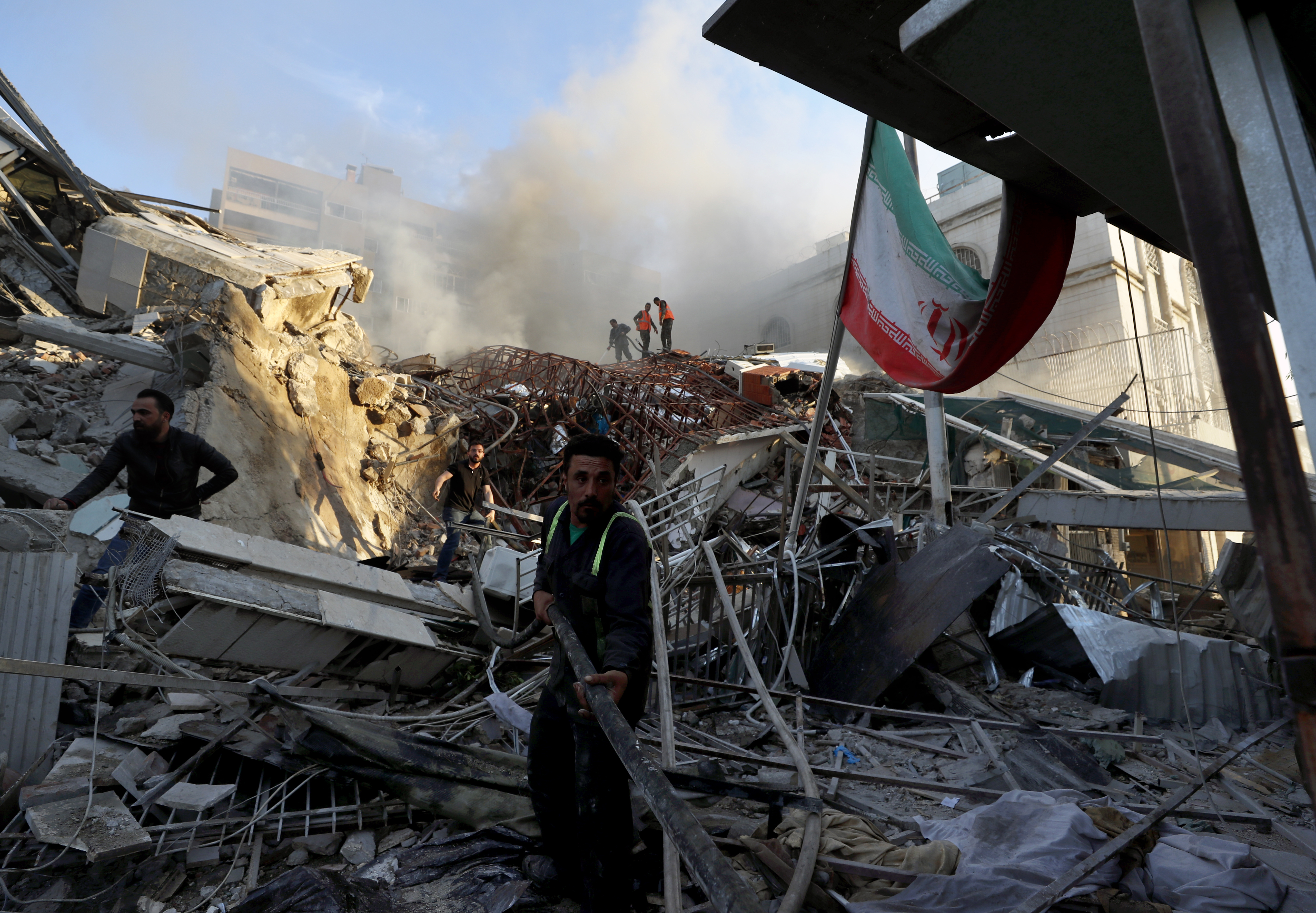 Rescue workers at the scene of the Iran consulate attack in Damascus. The building has been reduced to rubble. There is an Iranian flag to the right.