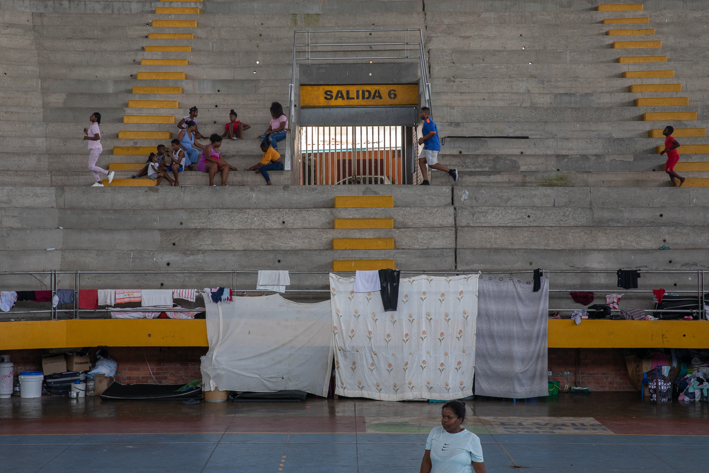 Inside a cement coliseum, families sit in the stands or set up bedsheets as walls for makeshift shelters.