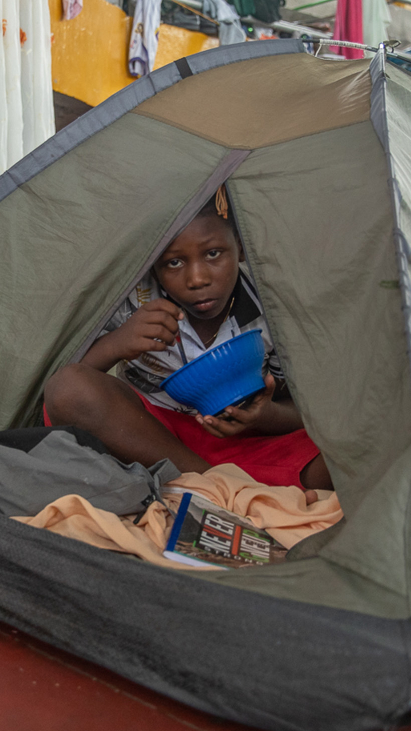A child eating from a bowl peers out from a tent.