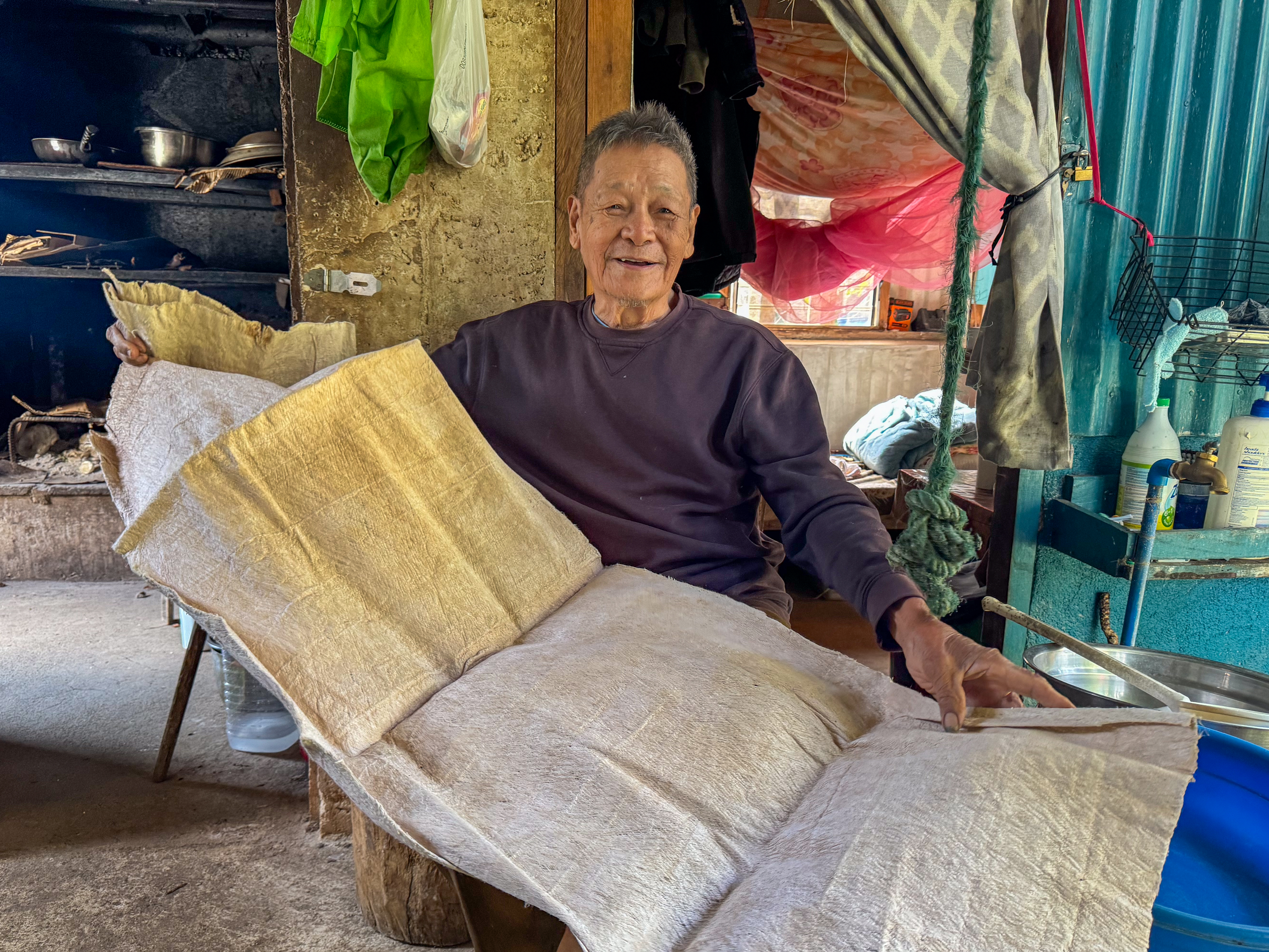 Eleuterio Malupa showing a sheath of tree bark. He is sitting in his home, The tree bark is pale coloured and has been folded.