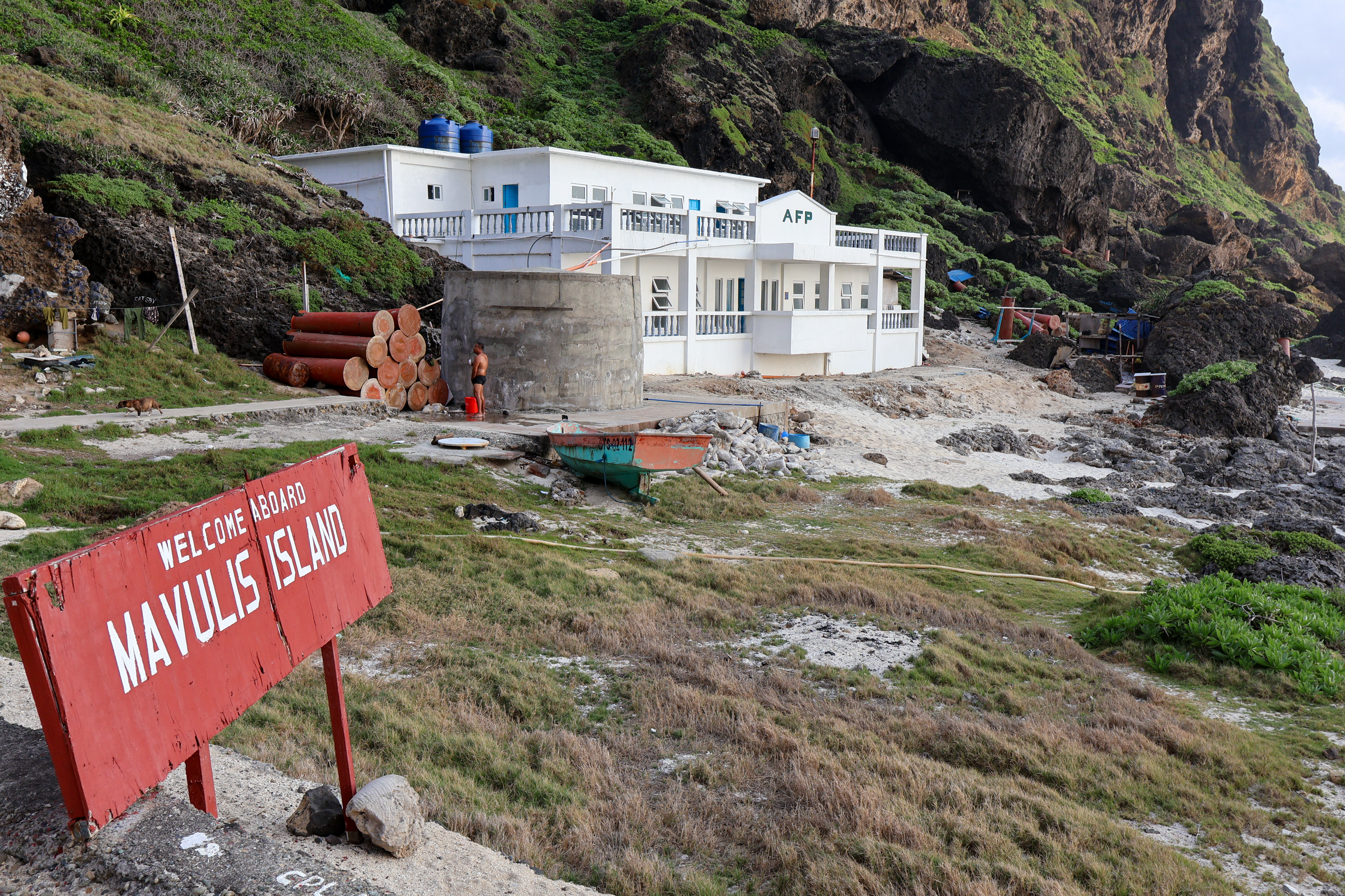 A view of the two storey barracks building on Mavulis. There are steep cliffs behind. A sign to the left says 'Welcome aboard Mavulis Island'