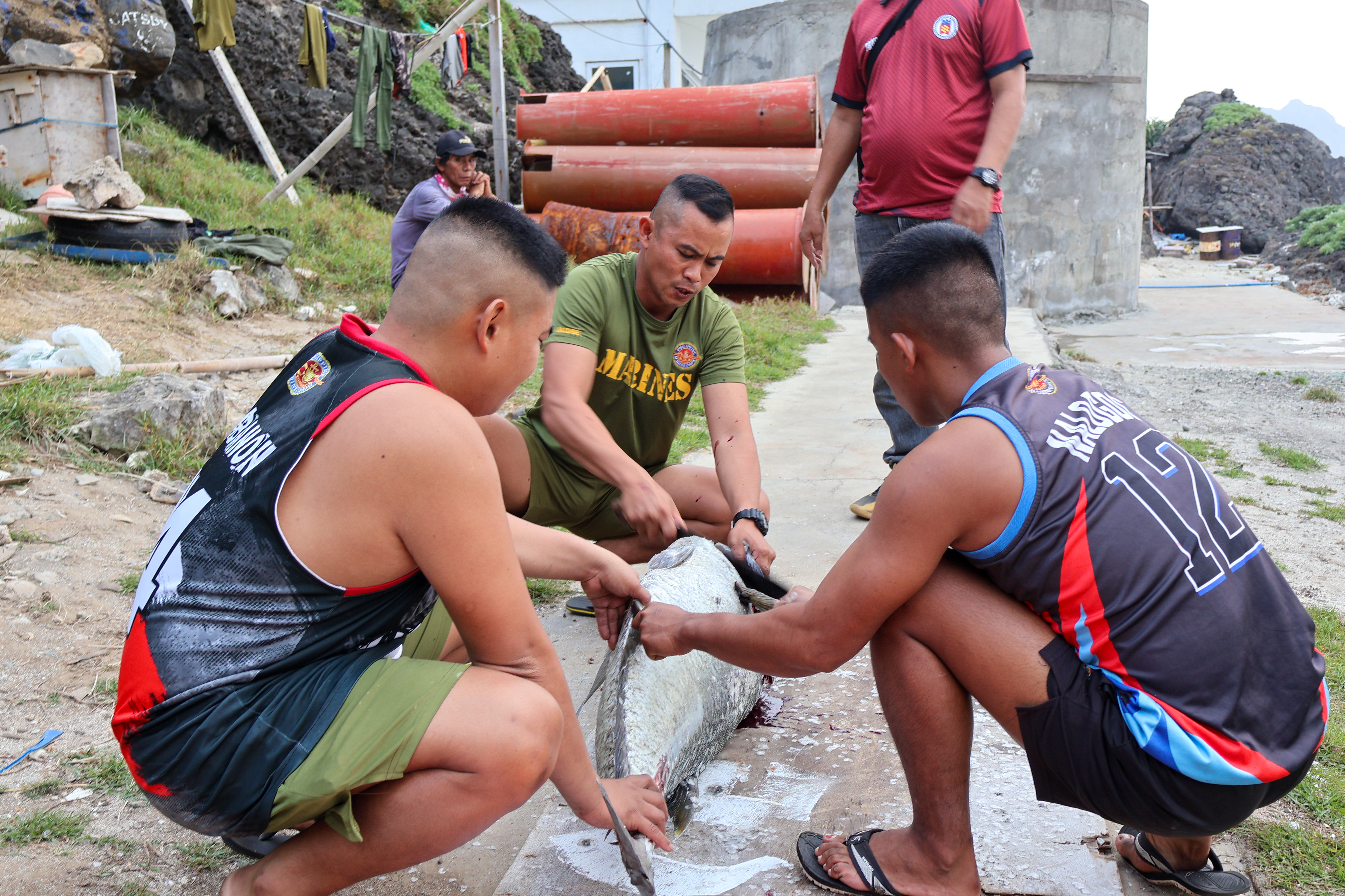Soldiers prepare a fish to make kinilaw. here are three of them squatted around it. One man is standing behind. The fish is quite large.