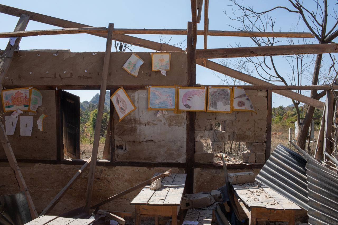 The ruins of the school in Daw Si Ei. The roof is missing and most of the wooden walls have collapsed. One remains standing, It has some of the children's work hung on it. There are ruined desks in front.