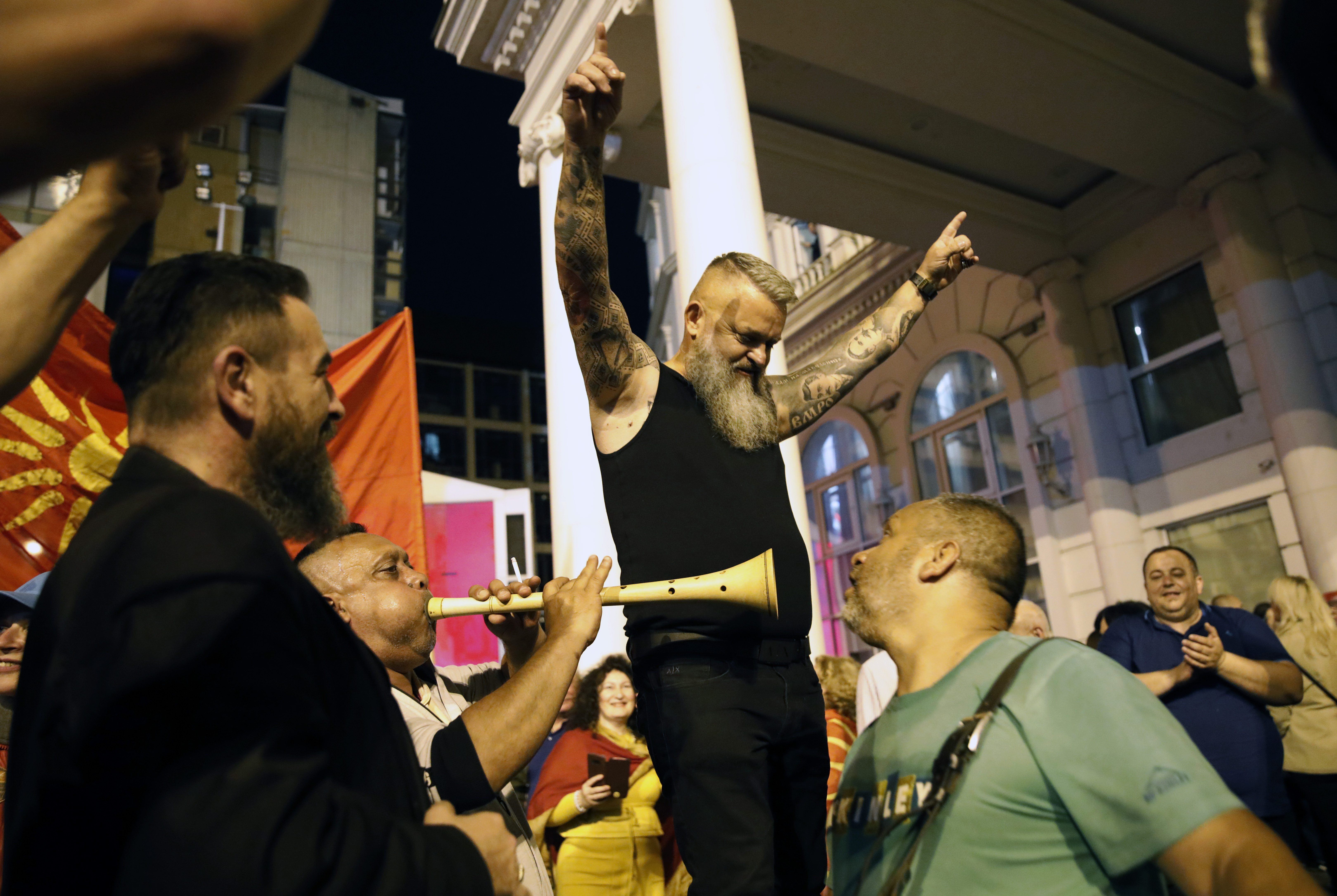 A bearded man in a black tank top raises both hands in the air as he celebrates with VMRO-DPMNE supporters in the streets of North Macedonia's capital. One fellow supporter blows into a horn-like instrument.