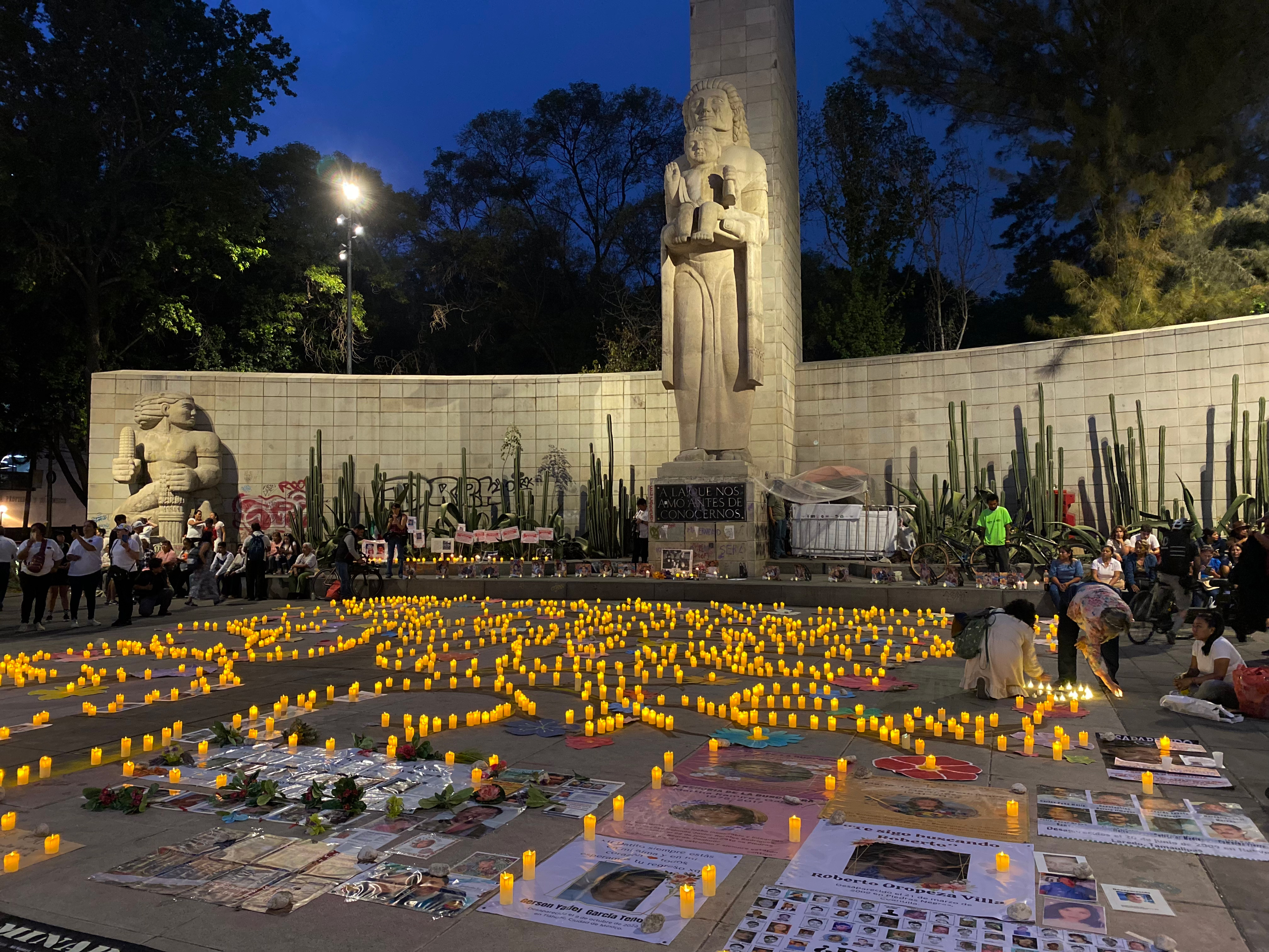 A photo of the Mother's Monument in Mexico City, an obelisk with the statue of a mother holding a child at its base. In front of the monument, the floor is filled with photographs and candles, as families seek justice for their missing loved ones.