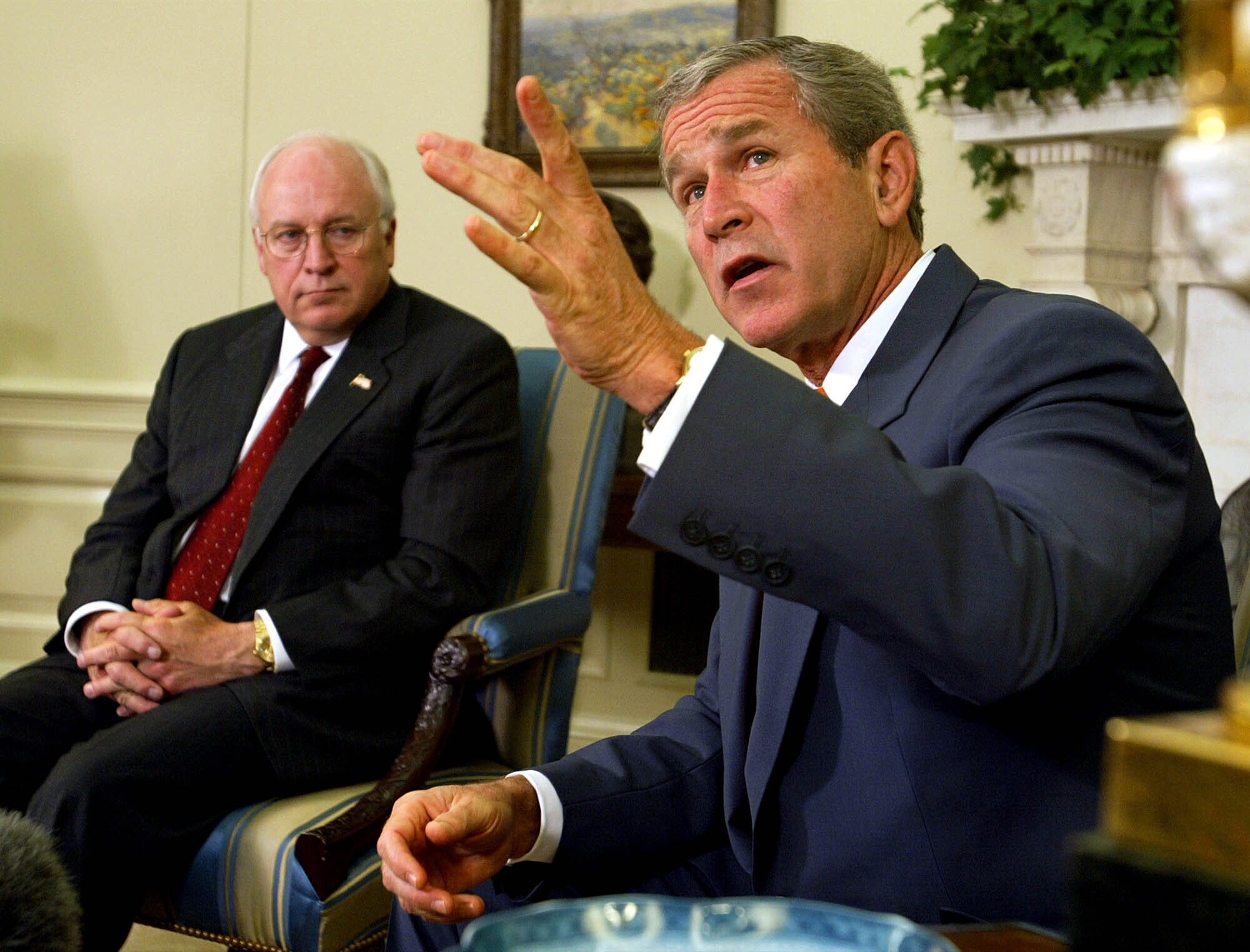 George W Bush gestures, seated, in the Oval Office. Behind him is Dick Cheney.